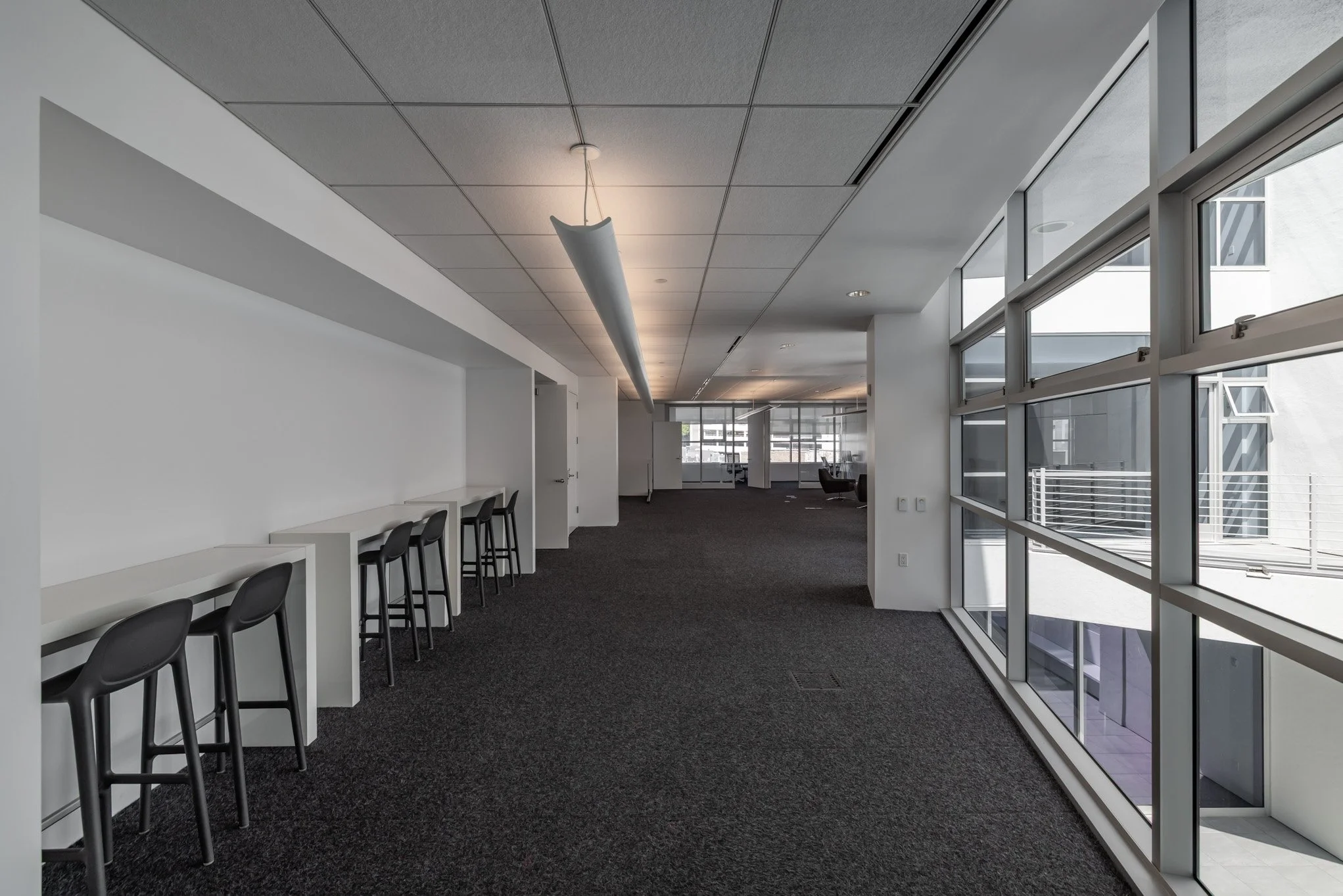 Empty modern office space with black chairs along a white counter, large windows, dark carpeted floor, and ceiling lights.