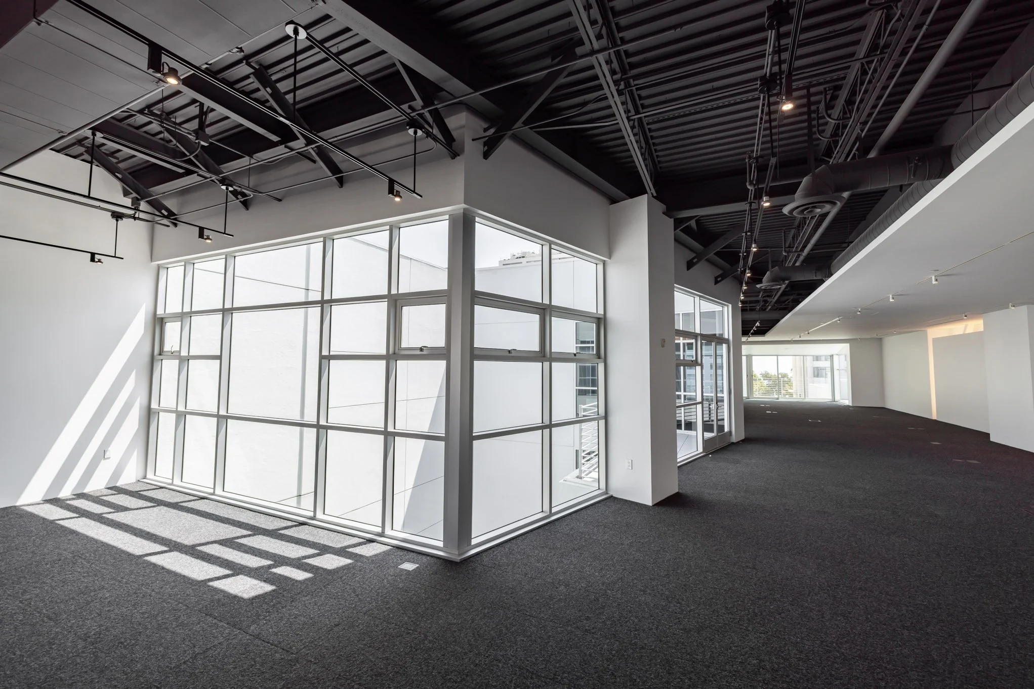 Empty modern office space with large windows, white walls, black ceiling with exposed fixtures, and dark carpeted floor.