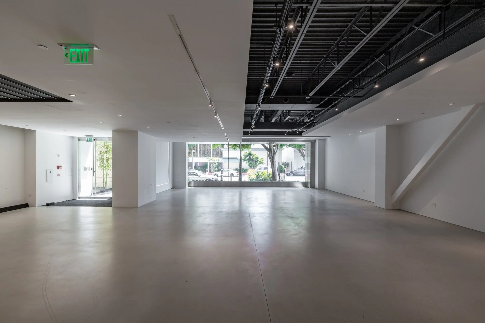 Empty interior space with large front window, white walls, and ceiling with exposed black structural elements and small ceiling lights.