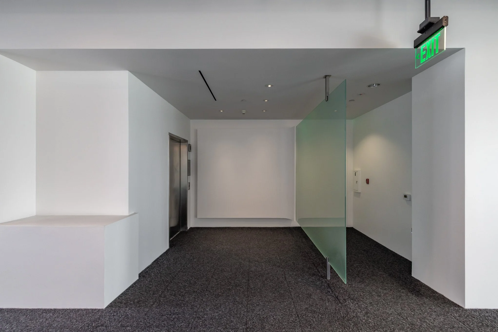 Empty modern hallway with white walls, black carpet, elevator, a green-lit emergency exit sign, and a glass panel divider.