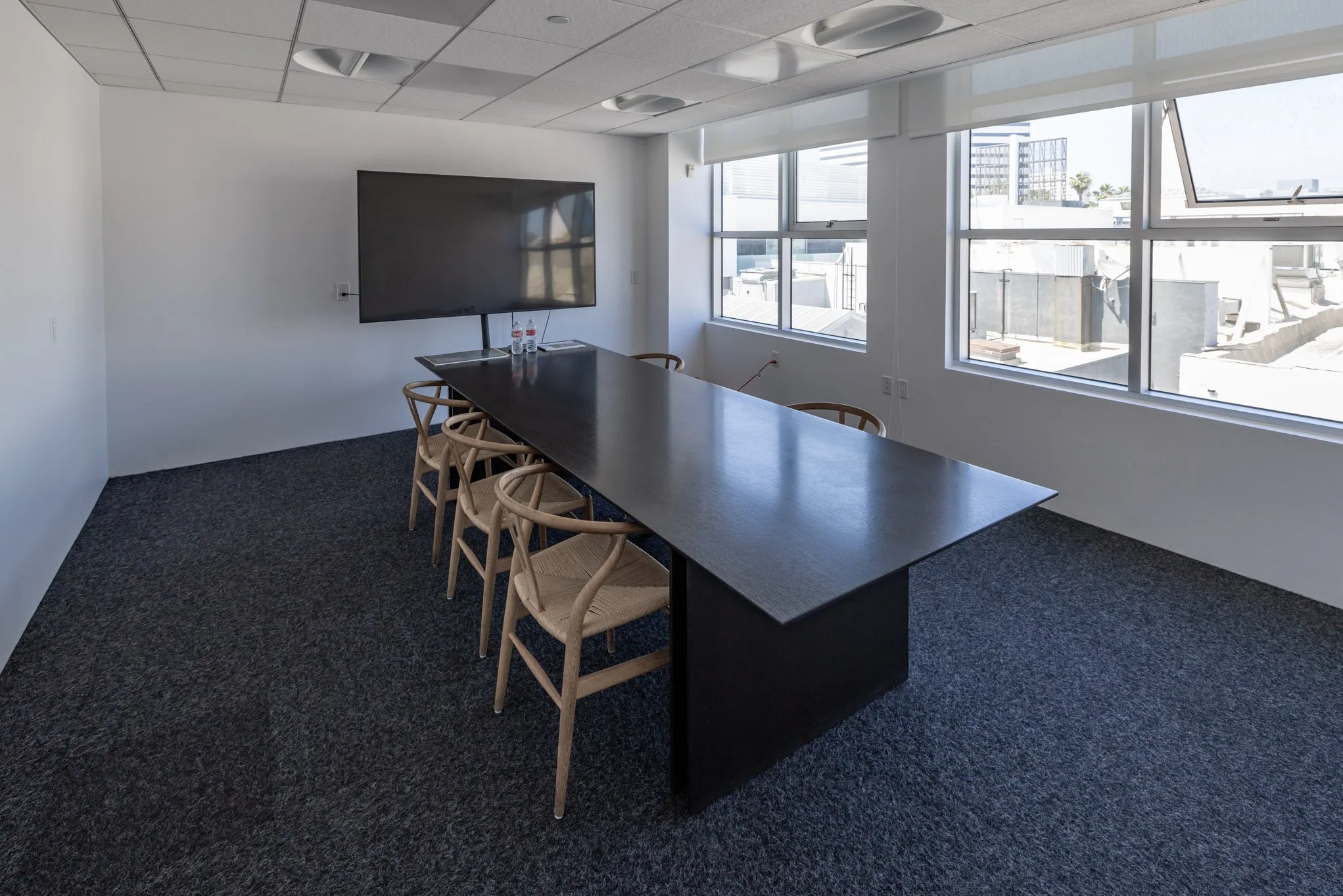 Empty conference room with a long black table, chairs, a large flat-screen TV on the wall, and floor-to-ceiling windows showing a cityscape.