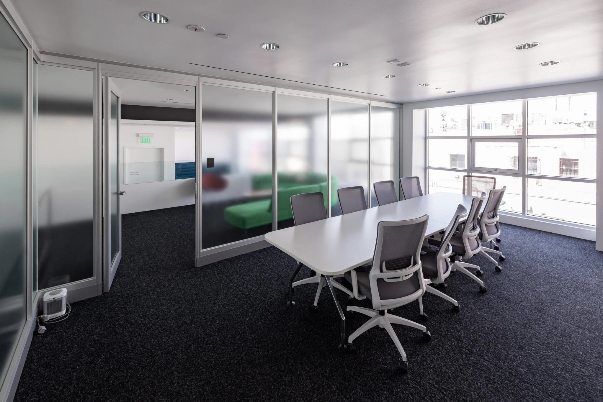 Empty conference room with long white table, nine office chairs, large windows, glass wall, and a green couch visible through the glass wall.