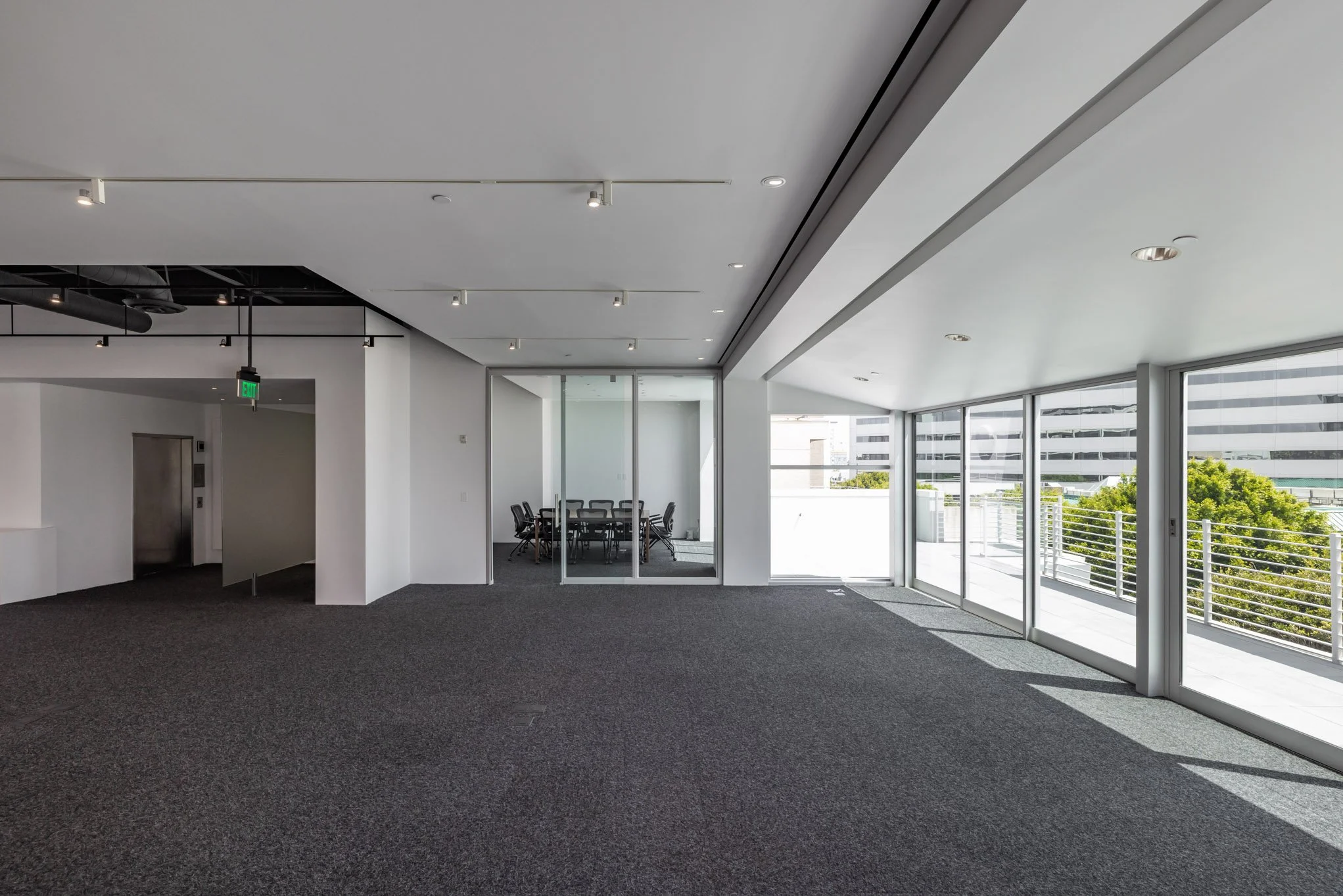 Empty office space with large windows, gray carpet, and a small conference room with a table and chairs.