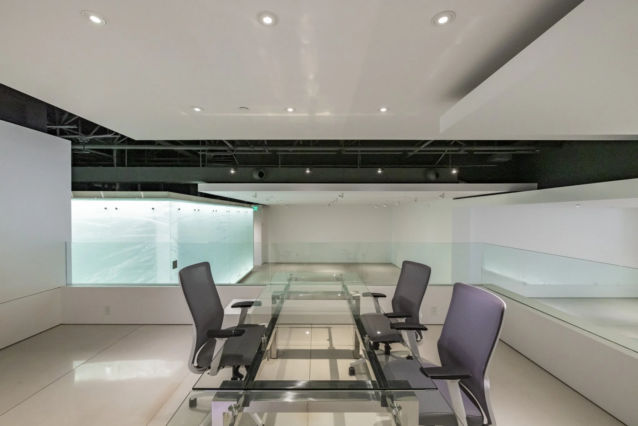 Empty modern conference room with glass table and gray chairs, illuminated by ceiling lights, with glass railing and wall with the ocean view in the background.