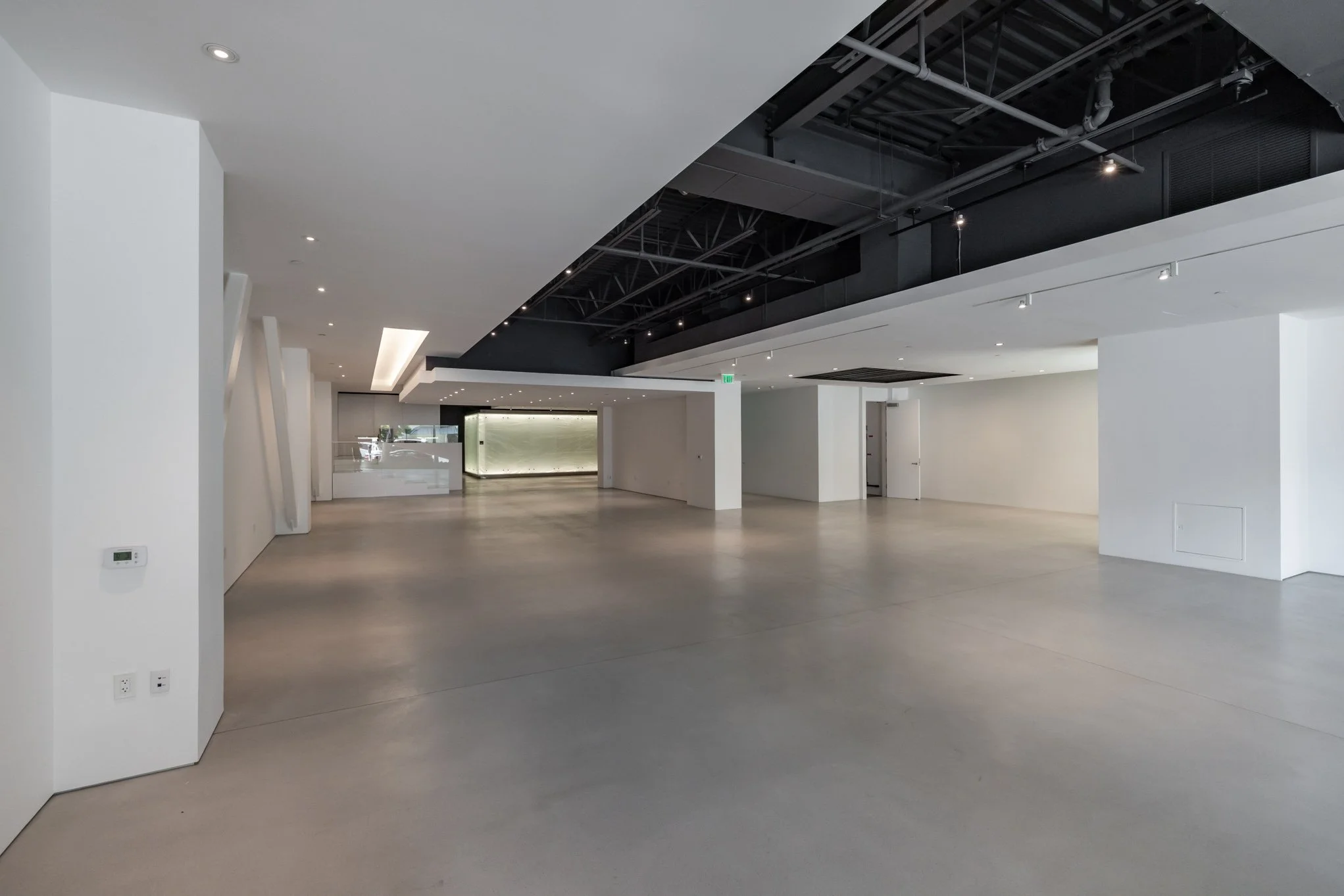 Empty modern interior space with white walls, gray concrete floors, and a black ceiling with exposed pipes and ductwork.