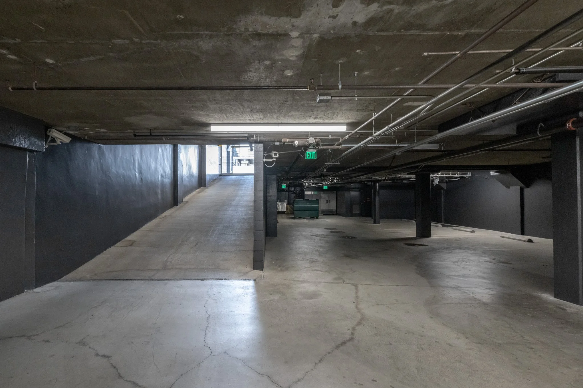 Underground parking garage with a ramp leading to the upper level, black walls, concrete floor, and exposed pipes on the ceiling.