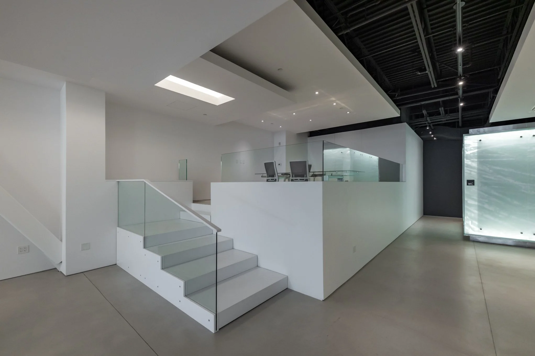 Empty modern office space with white walls, glass railing, staircase, and black ceiling with lights.