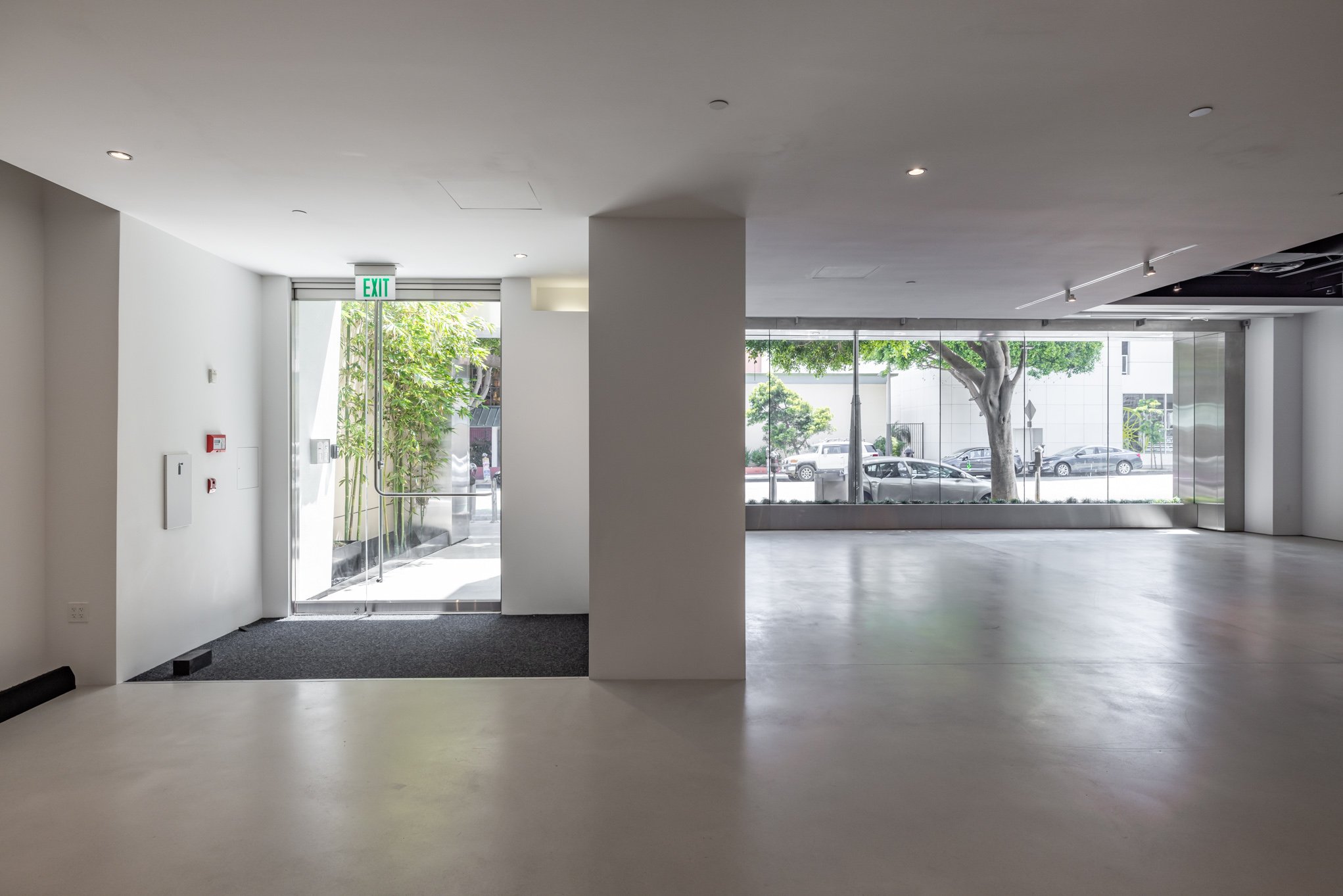 Empty interior space with large glass windows, door with EXIT sign, and trees outside.