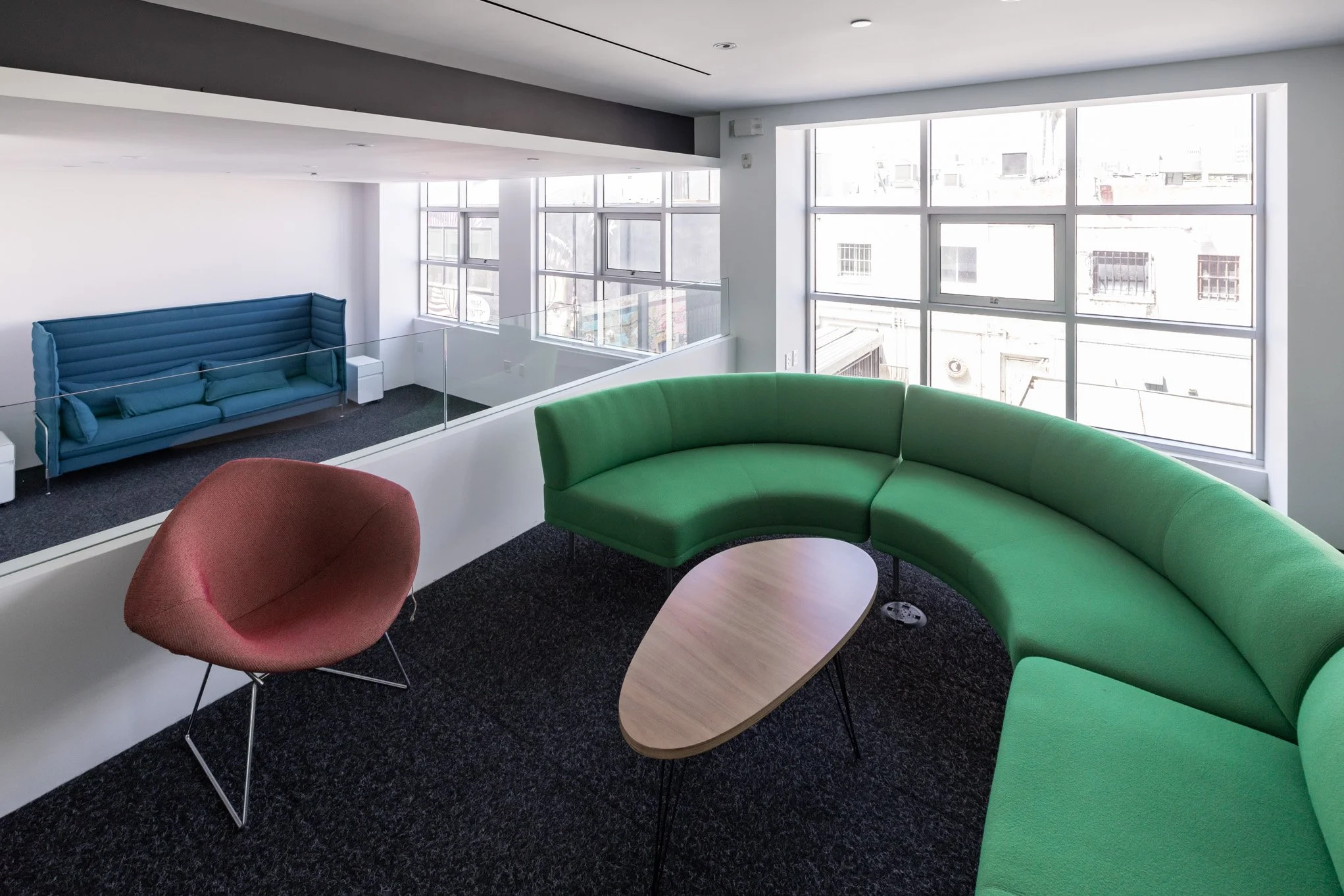 Modern office lounge area with large window, green curved sofa, pink chair, wooden coffee table, and blue sofa in the background.