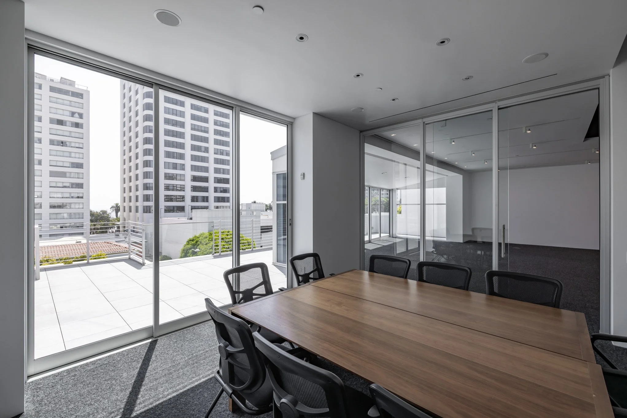 Modern conference room with large glass sliding doors leading to a balcony, view of high-rise buildings, wooden conference table, and black office chairs.