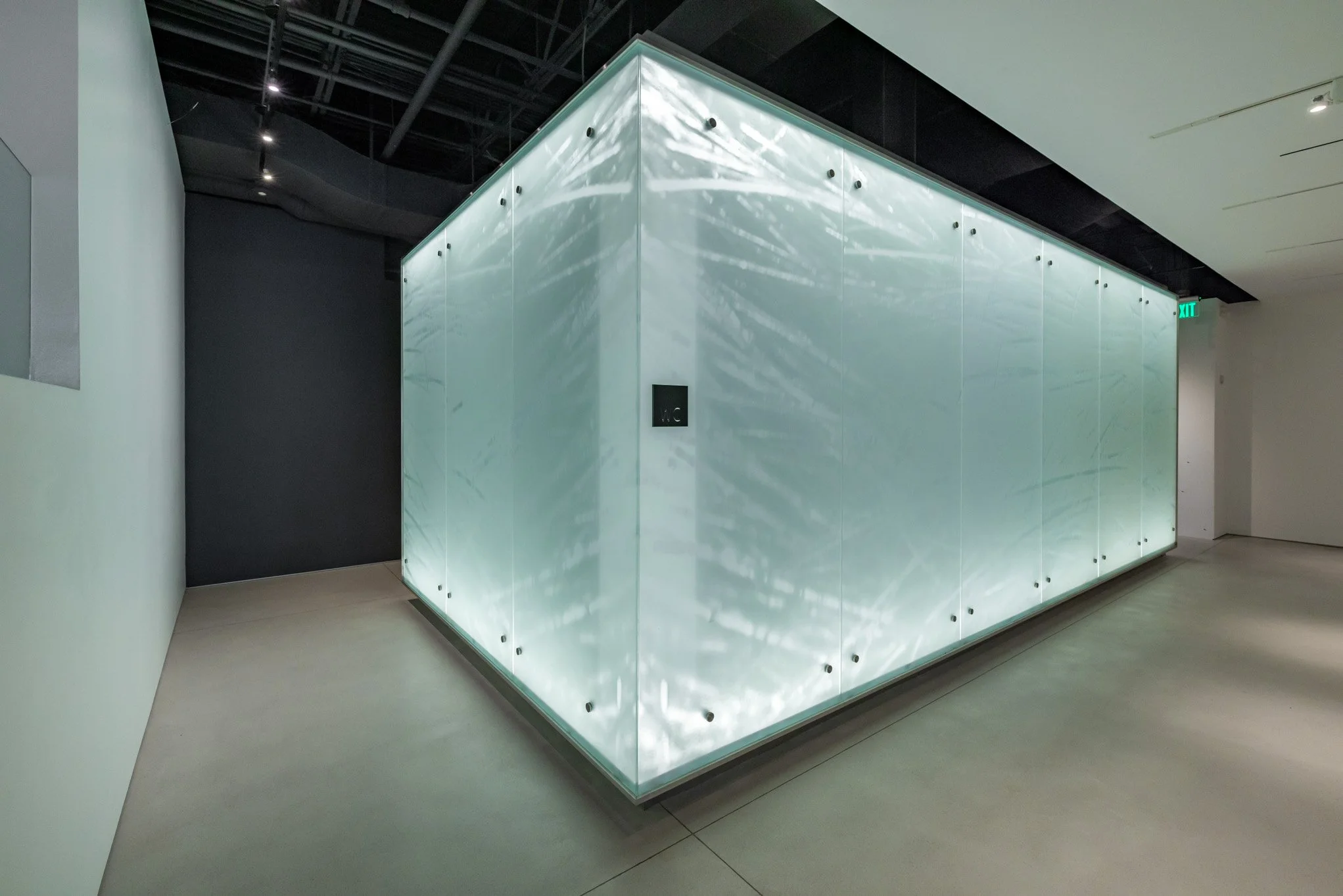 Illuminated glass cube with water-like light reflections in a modern interior space, with an exit sign and a dark corner.