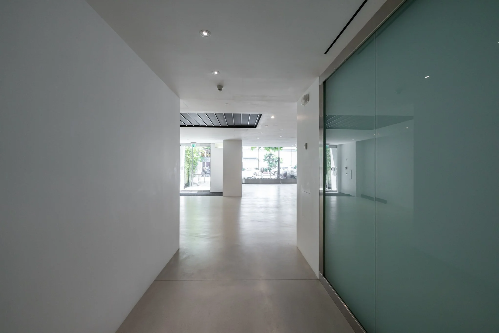 Empty modern lobby or hallway with white walls, light-colored flooring, ceiling with recessed lighting, glass wall on the right, and large windows and glass entrance doors at the end, allowing natural light to brighten the space.