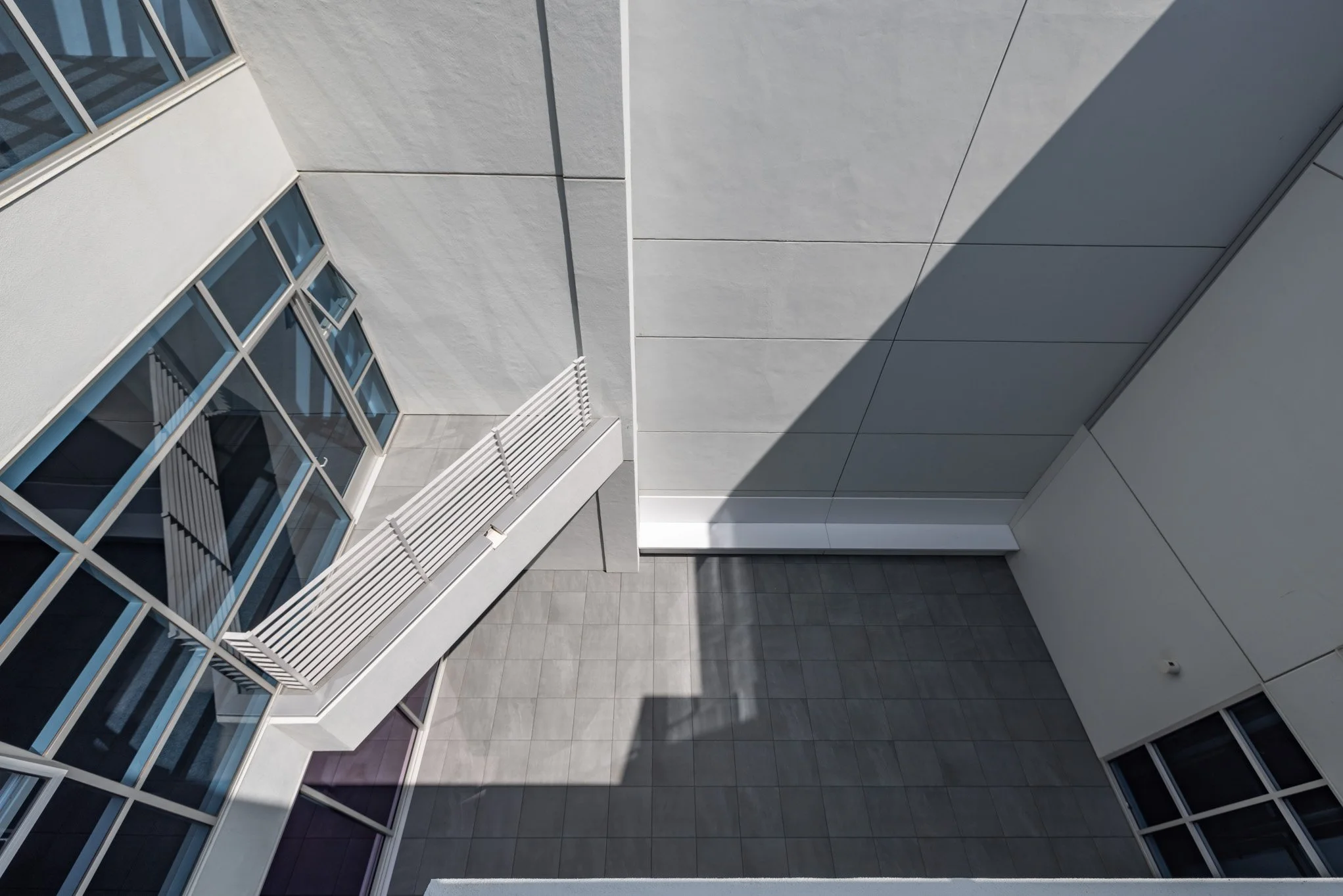 Looking up into a modern courtyard of a building with gray walls, windows, and a white balcony with a railing. Shadows cast by the building create geometric patterns on the surfaces.
