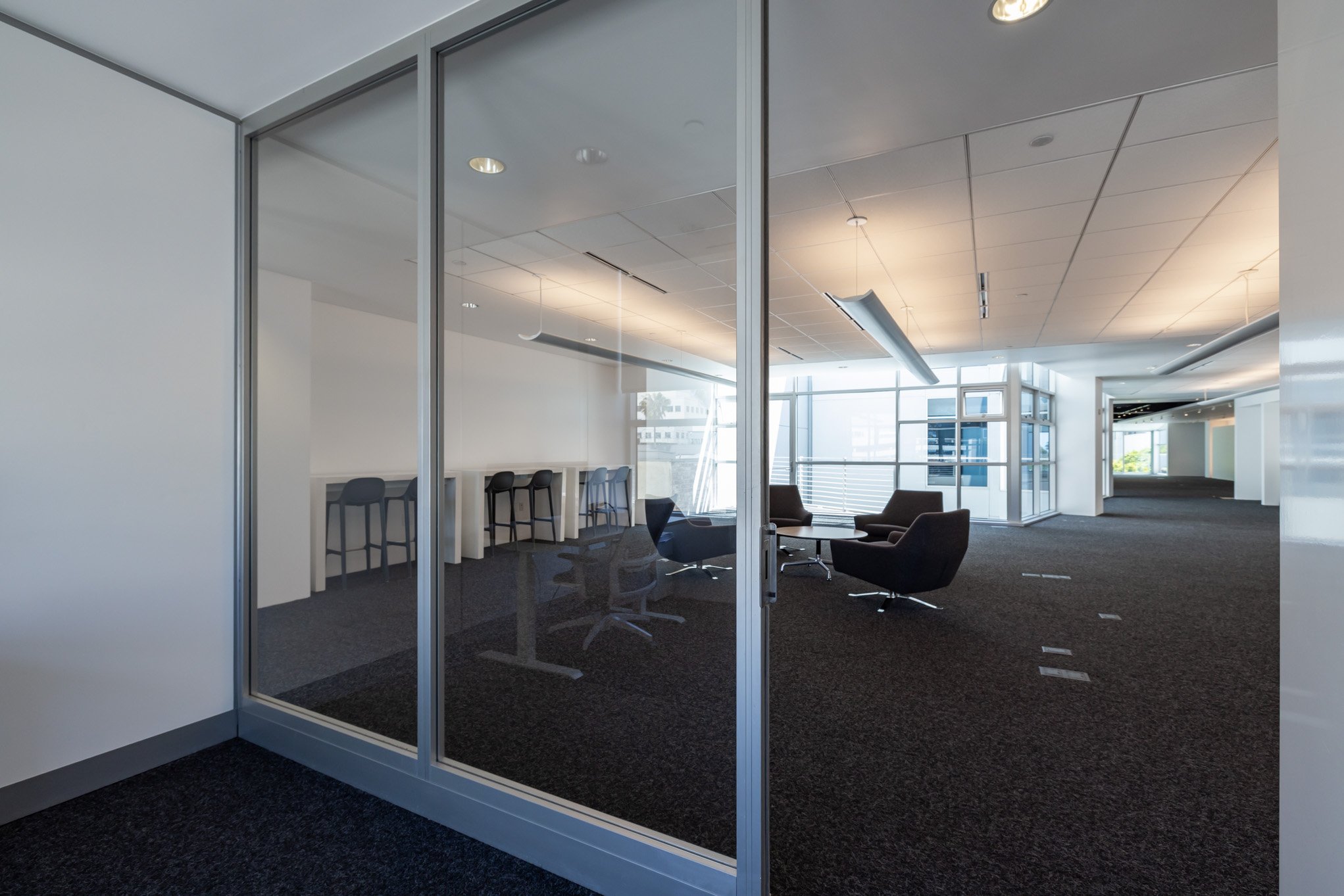 Empty modern office with glass walls, black lounge chairs, and high-top counter with chairs, illuminated by natural light from large windows.