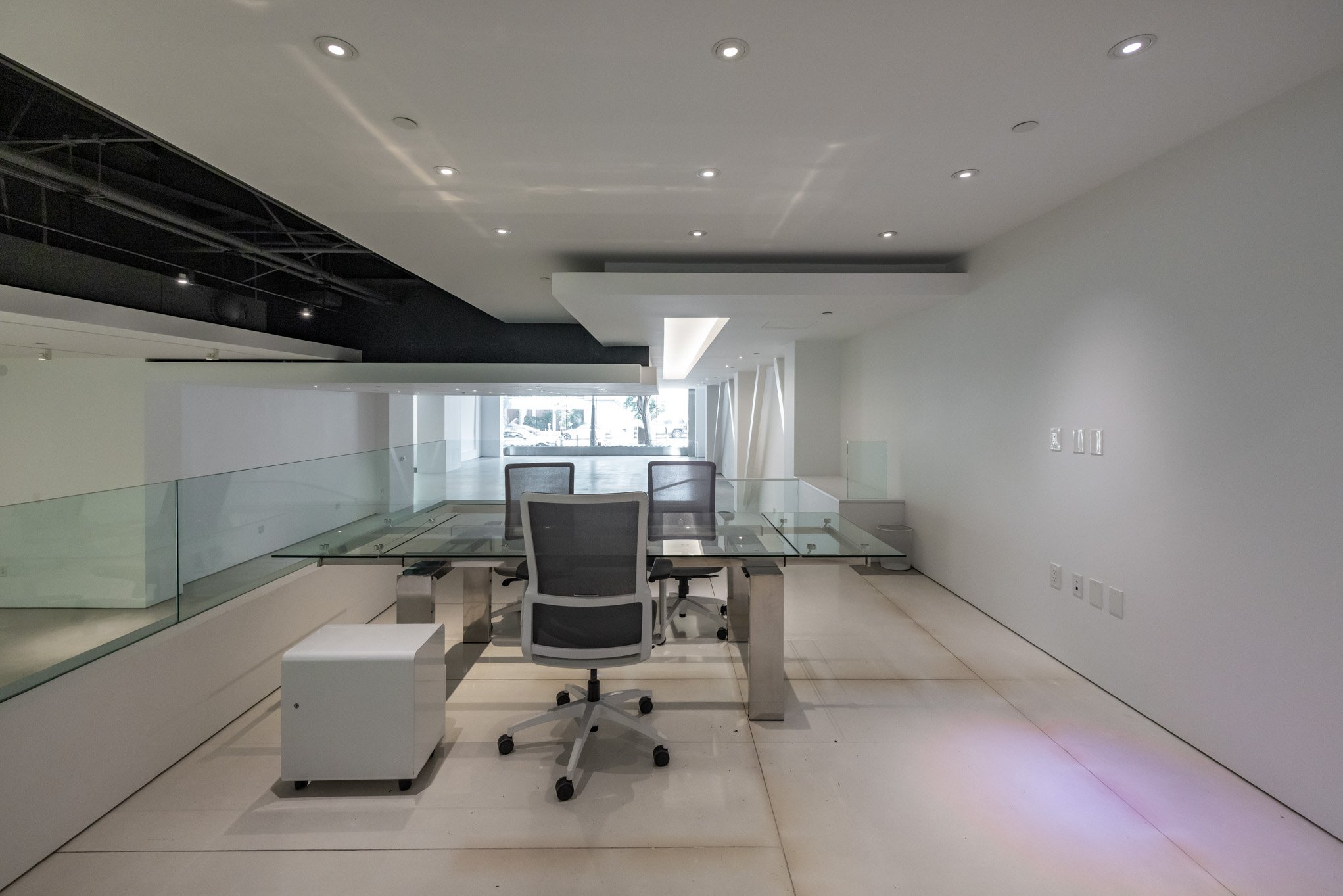 Empty modern office space with glass desk, office chairs, and white walls, minimal decor, and natural light coming through large windows.