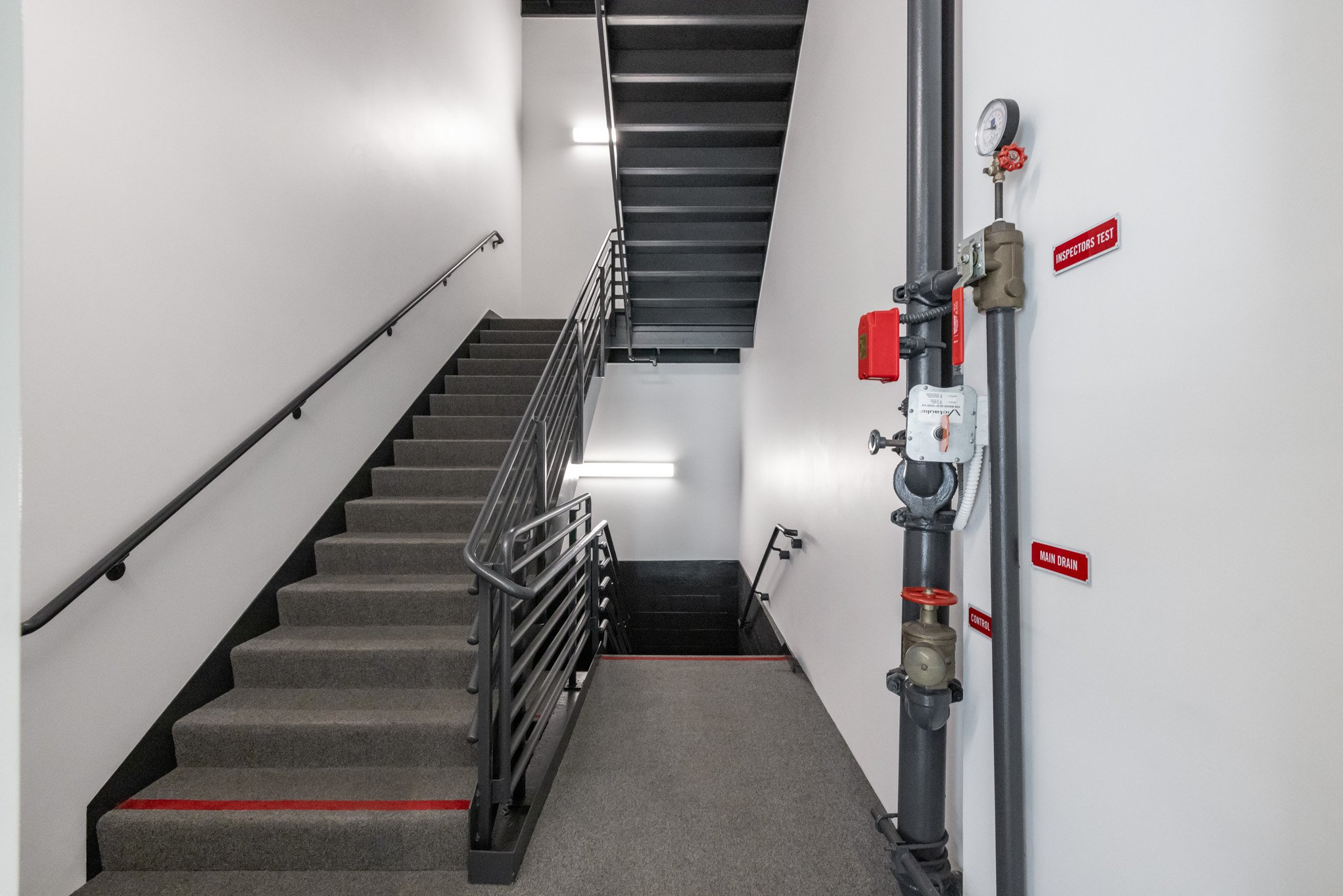 Indoor staircase with gray carpeted steps and metal handrails next to a white wall. Fire safety equipment and pipes are visible on the right side of the image.
