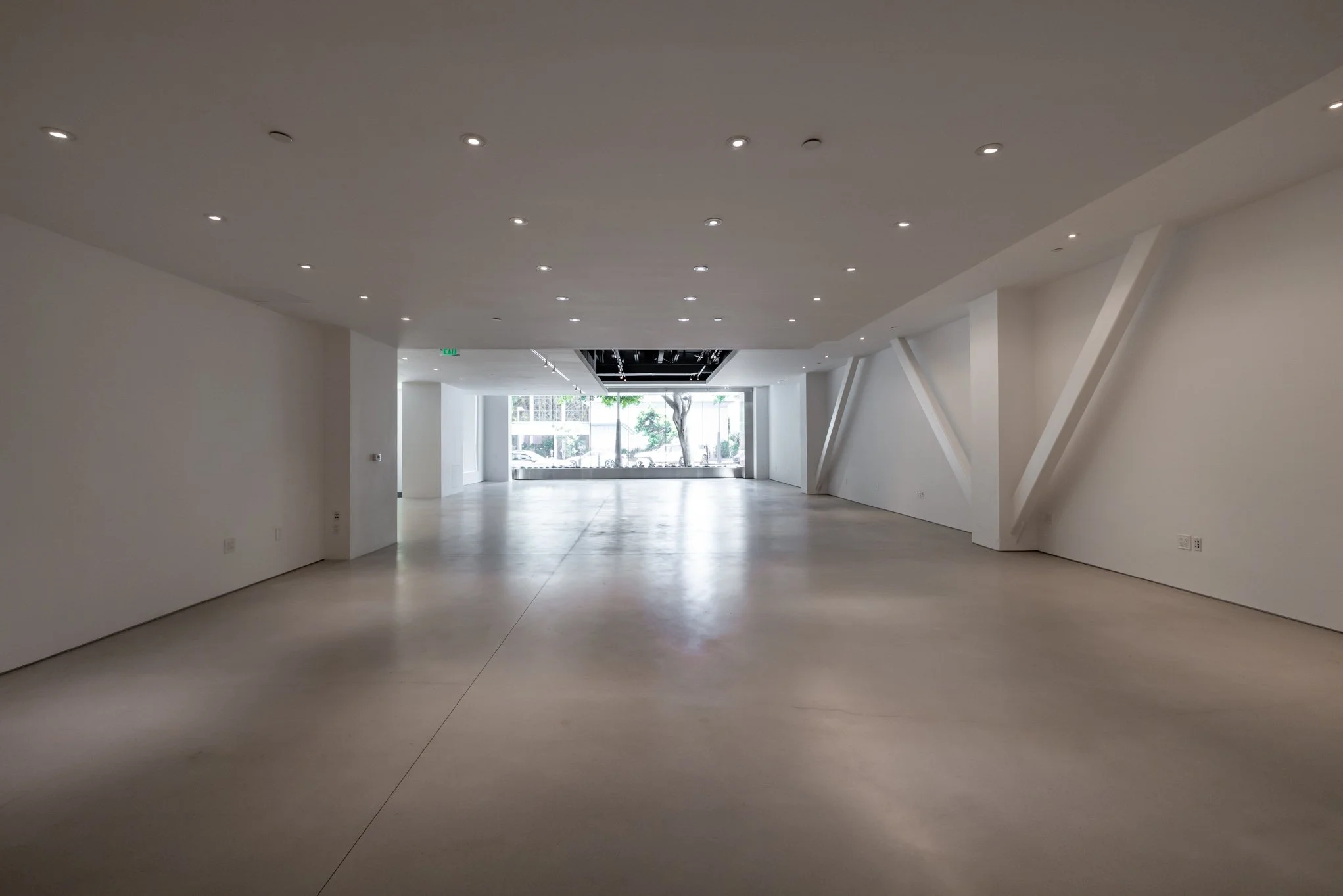 Empty commercial or gallery space with white walls, polished concrete floor, and recessed ceiling lights, with front glass doors and windows showing a view outside.