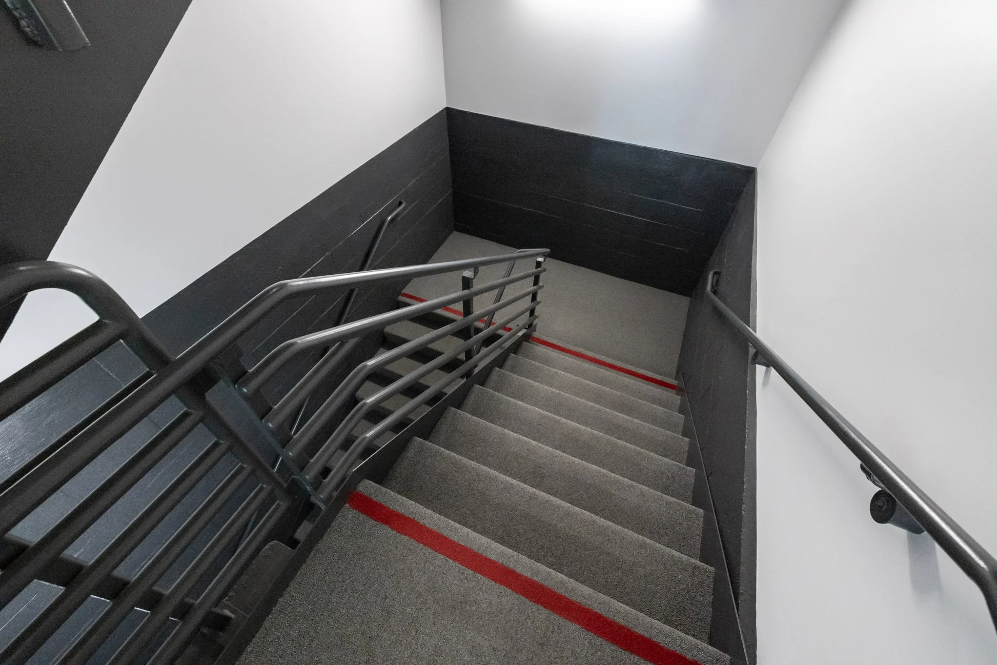 Indoor staircase with gray carpeted steps, black metal railing, and black wall panels, viewed from the top.