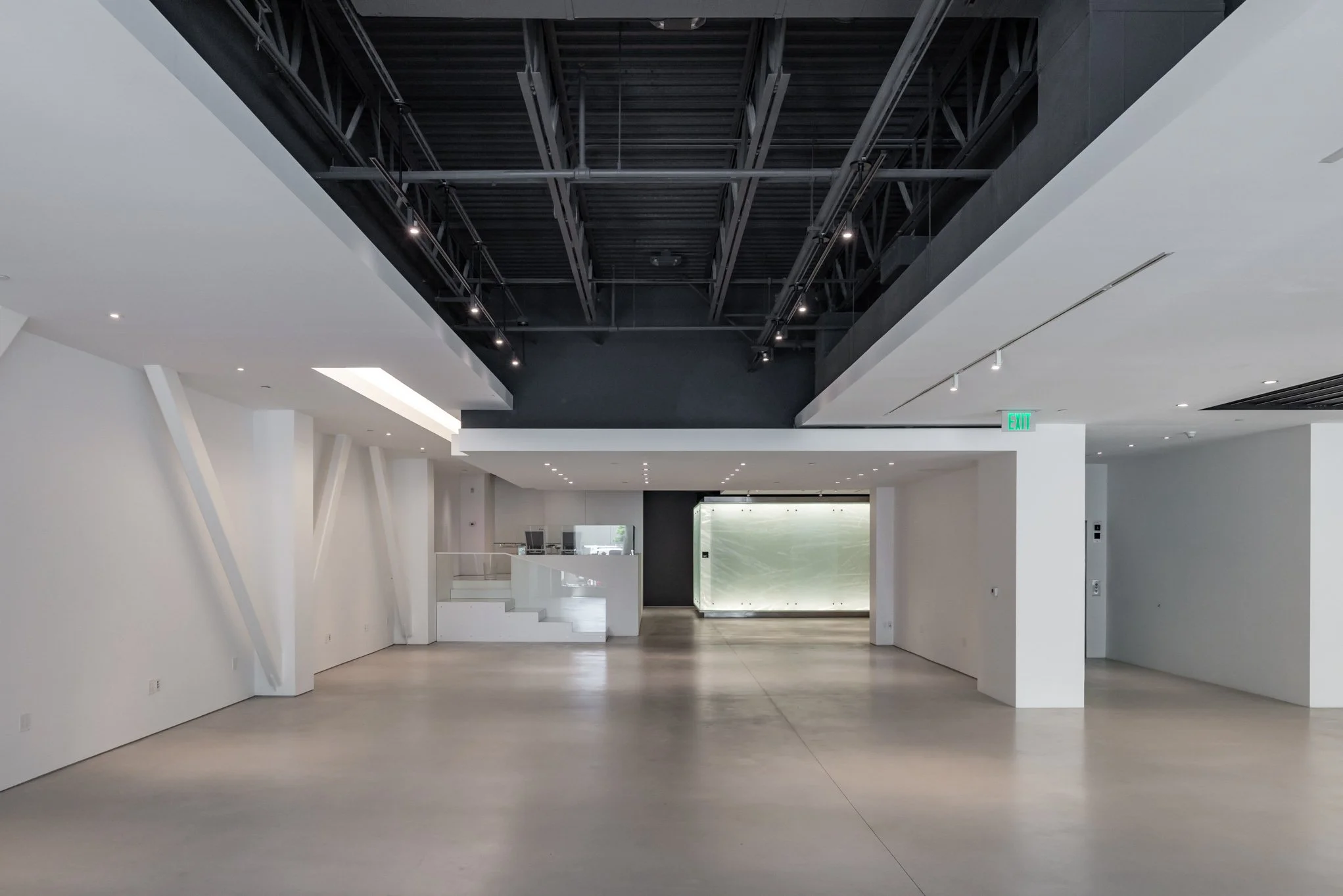 Empty modern interior space with white walls, polished concrete floors, black ceiling with exposed pipes, and illuminated reception desk in the background.