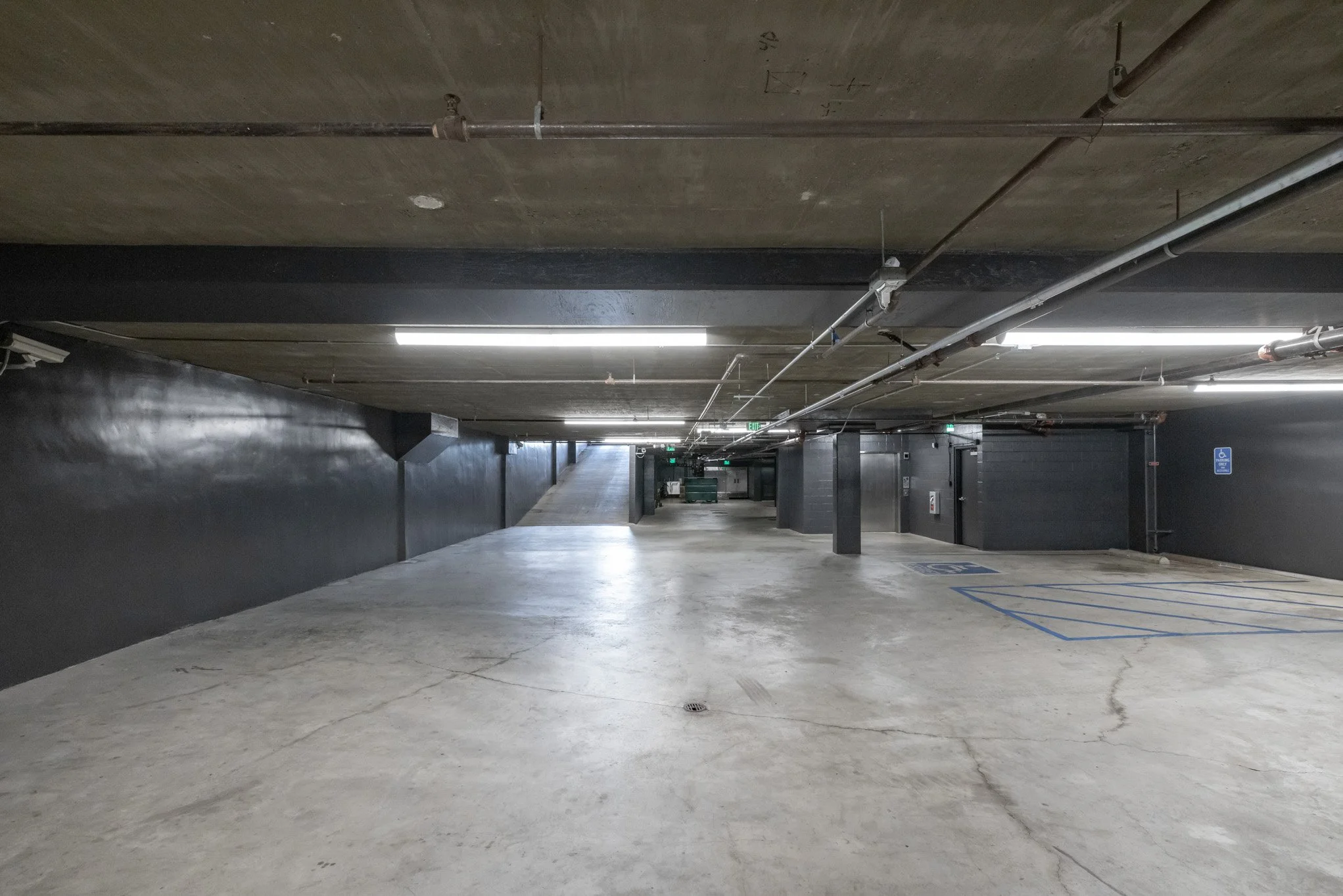 Underground parking garage with concrete floor and ceiling, black walls, pipes and fluorescent lights, including a reserved handicapped parking space.