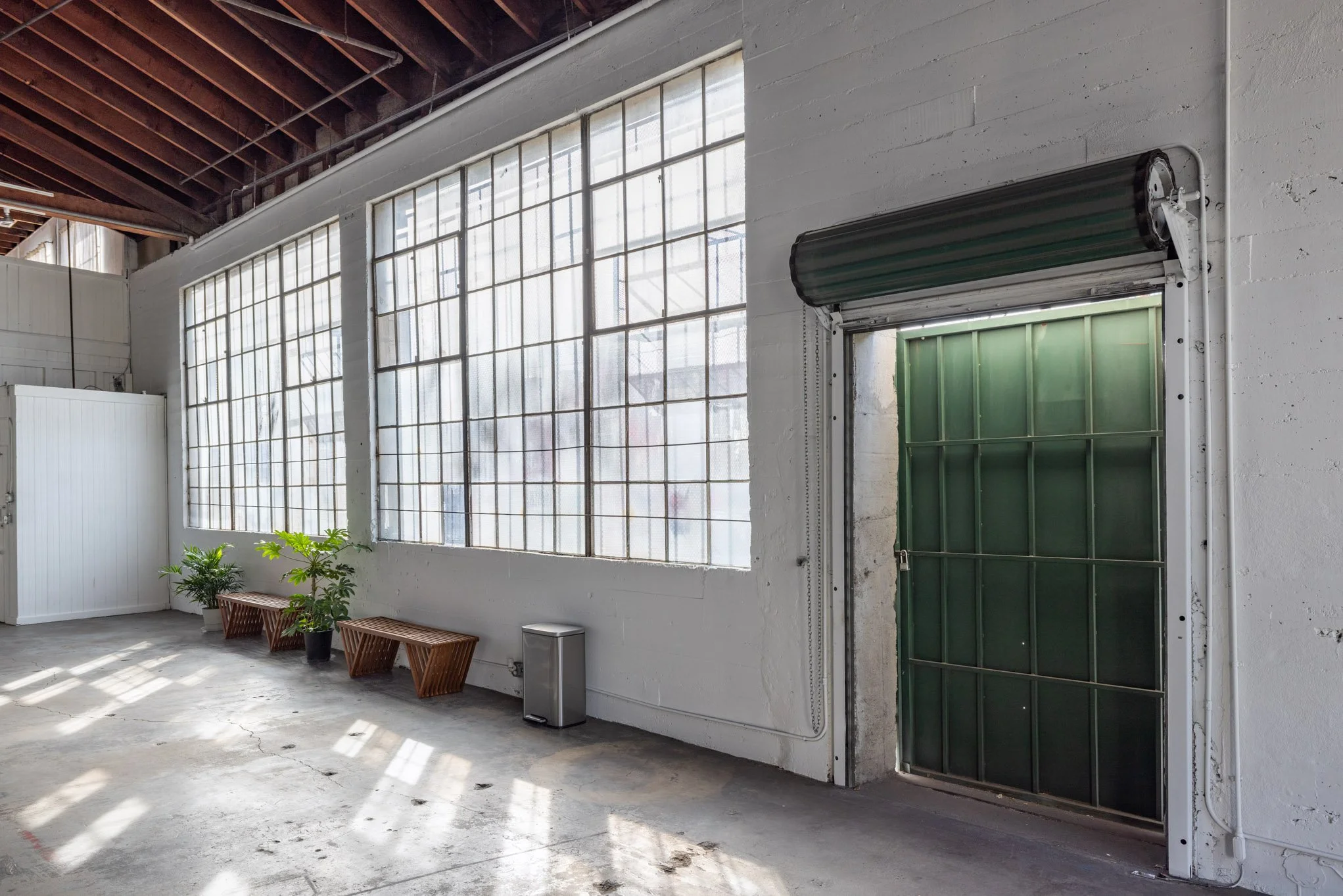 Industrial interior with large windows, potted plants, wooden benches, and a green rolling door.