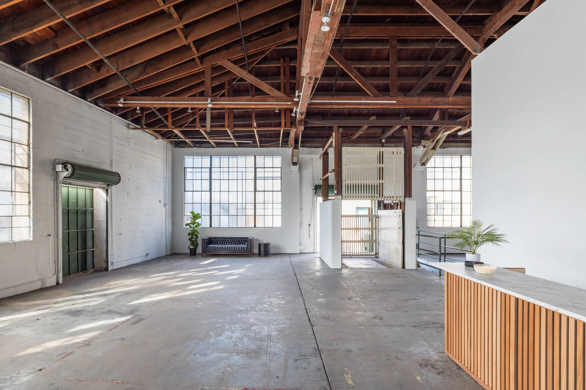 Large open industrial-style space with high wooden ceiling, large windows, minimal furniture, concrete floor, and a reception desk with a small potted plant.