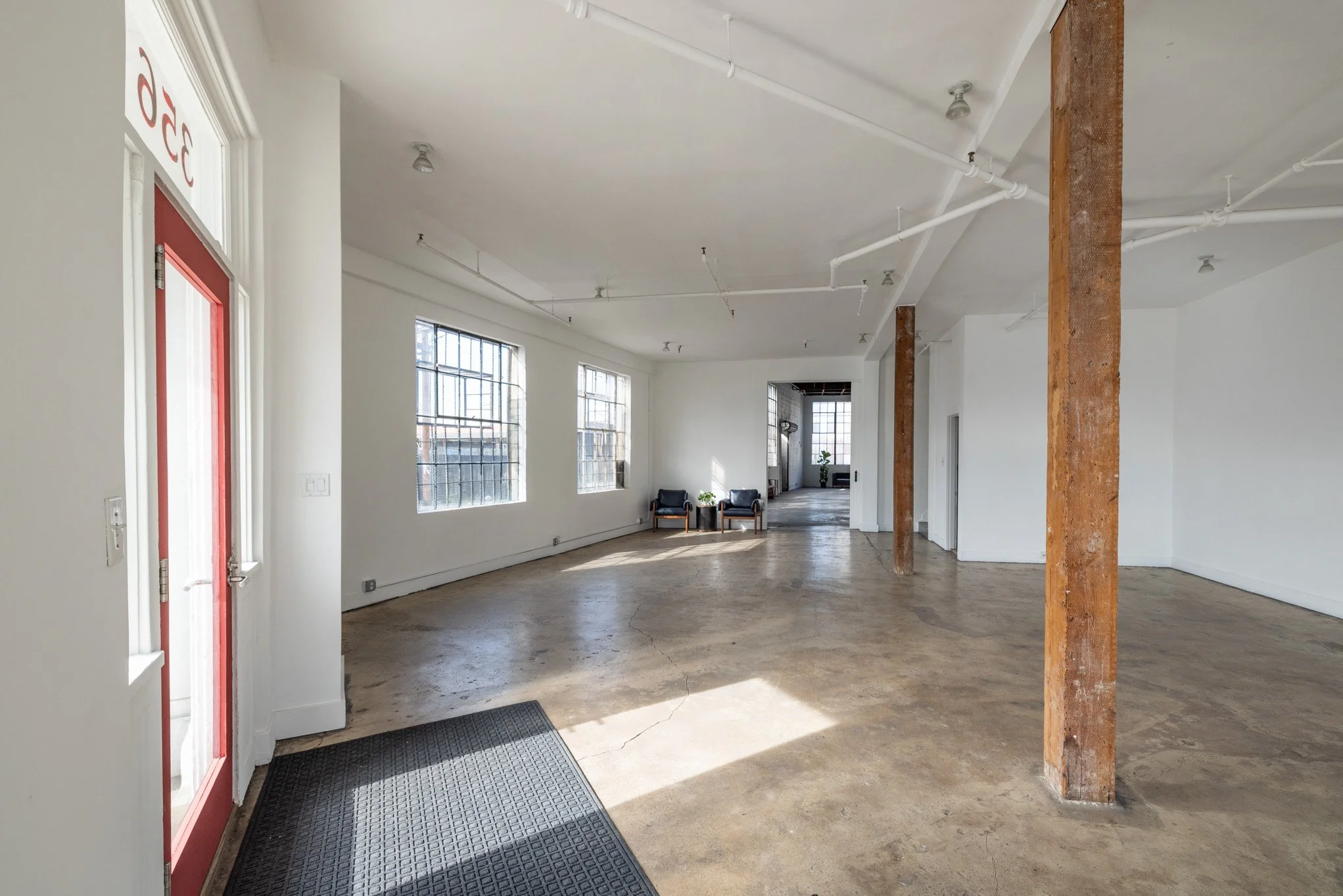 Empty commercial space with large windows, wooden support beams, concrete floors, and a corner seating area with two black chairs and a small table.