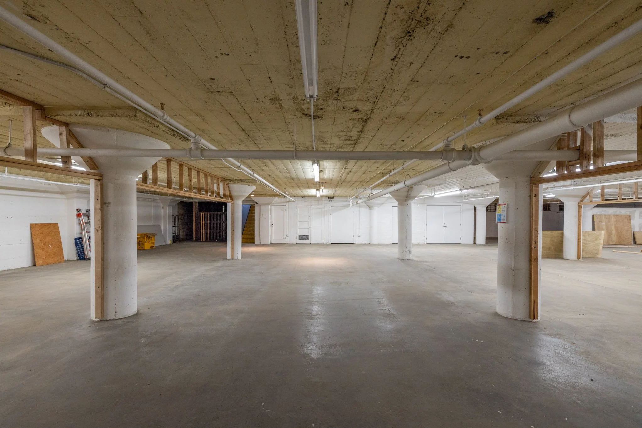 Empty parking garage with concrete floors, white painted walls, exposed pipes, and concrete support columns.