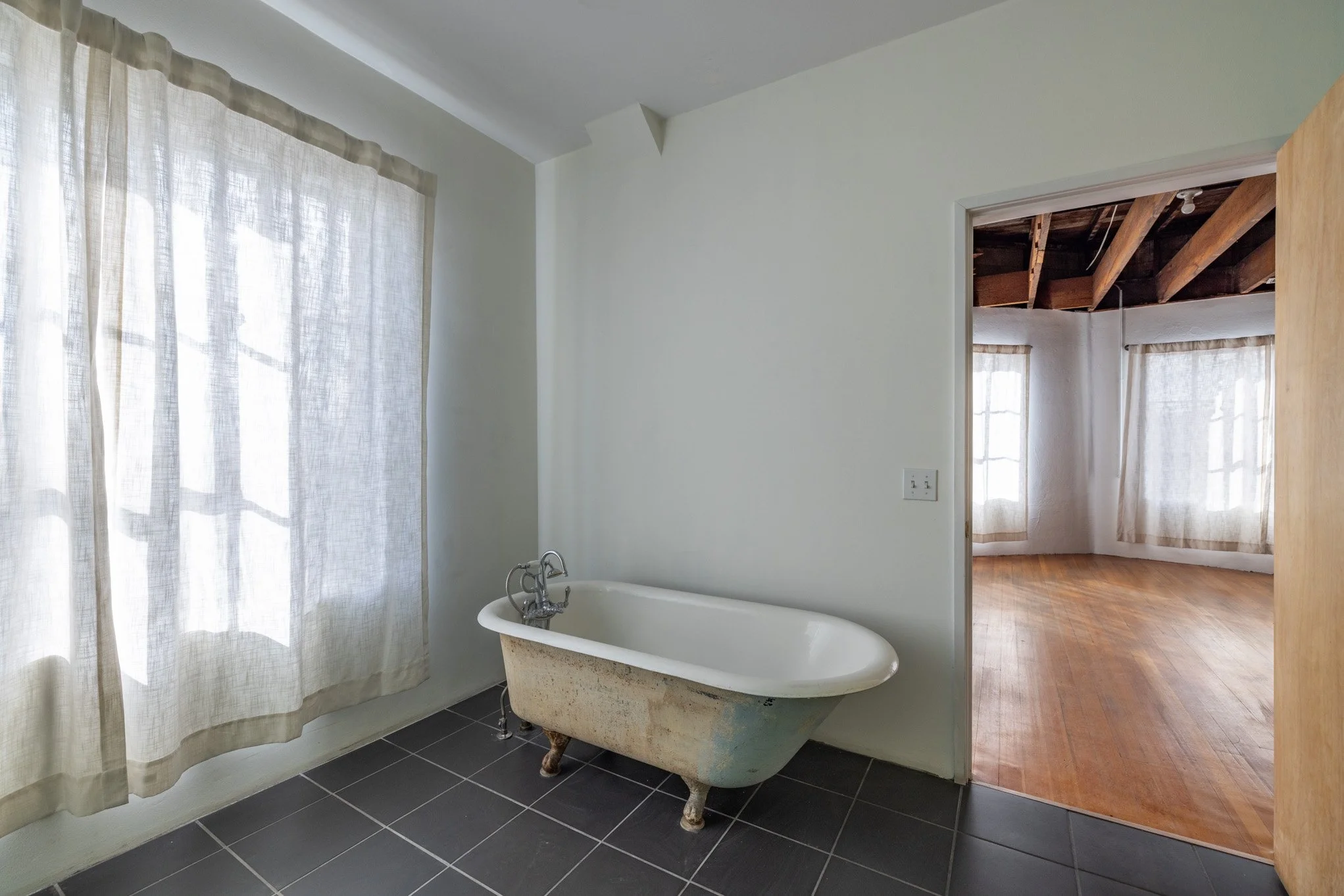 A vintage clawfoot bathtub is situated in a room with light-colored walls, a window with sheer curtains, and dark tiled flooring.