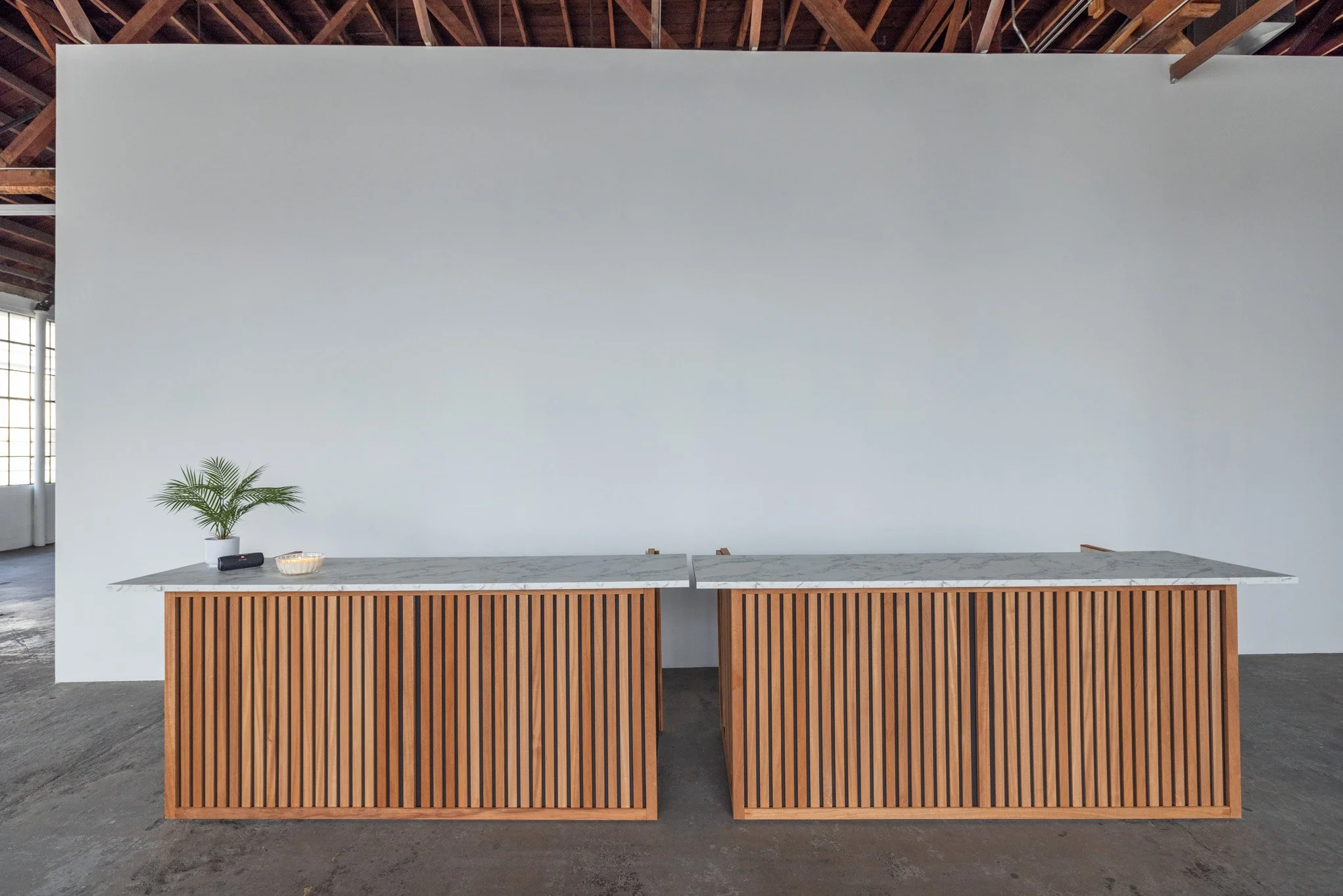 Two modern kitchen islands with marble countertops and wooden slatted fronts are placed in front of a plain white wall in an industrial-style room with exposed ceiling beams and large windows.
