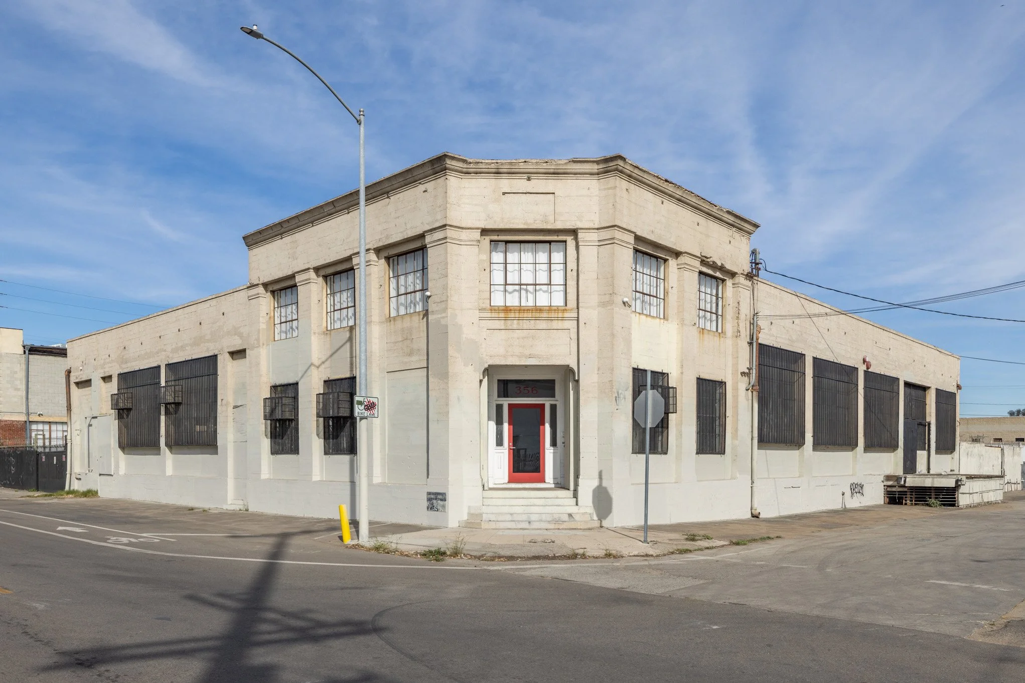 A beige, two-story corner building with barred windows and a red door, situated on a paved street under a blue sky.