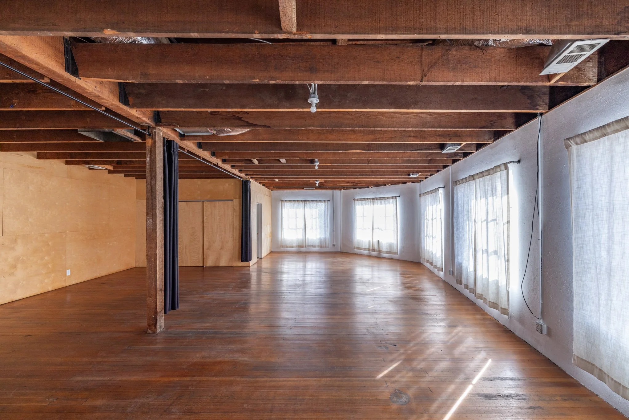Empty room with wooden floors, bare ceiling with exposed beams, and large windows with curtains allowing sunlight to fill the space.