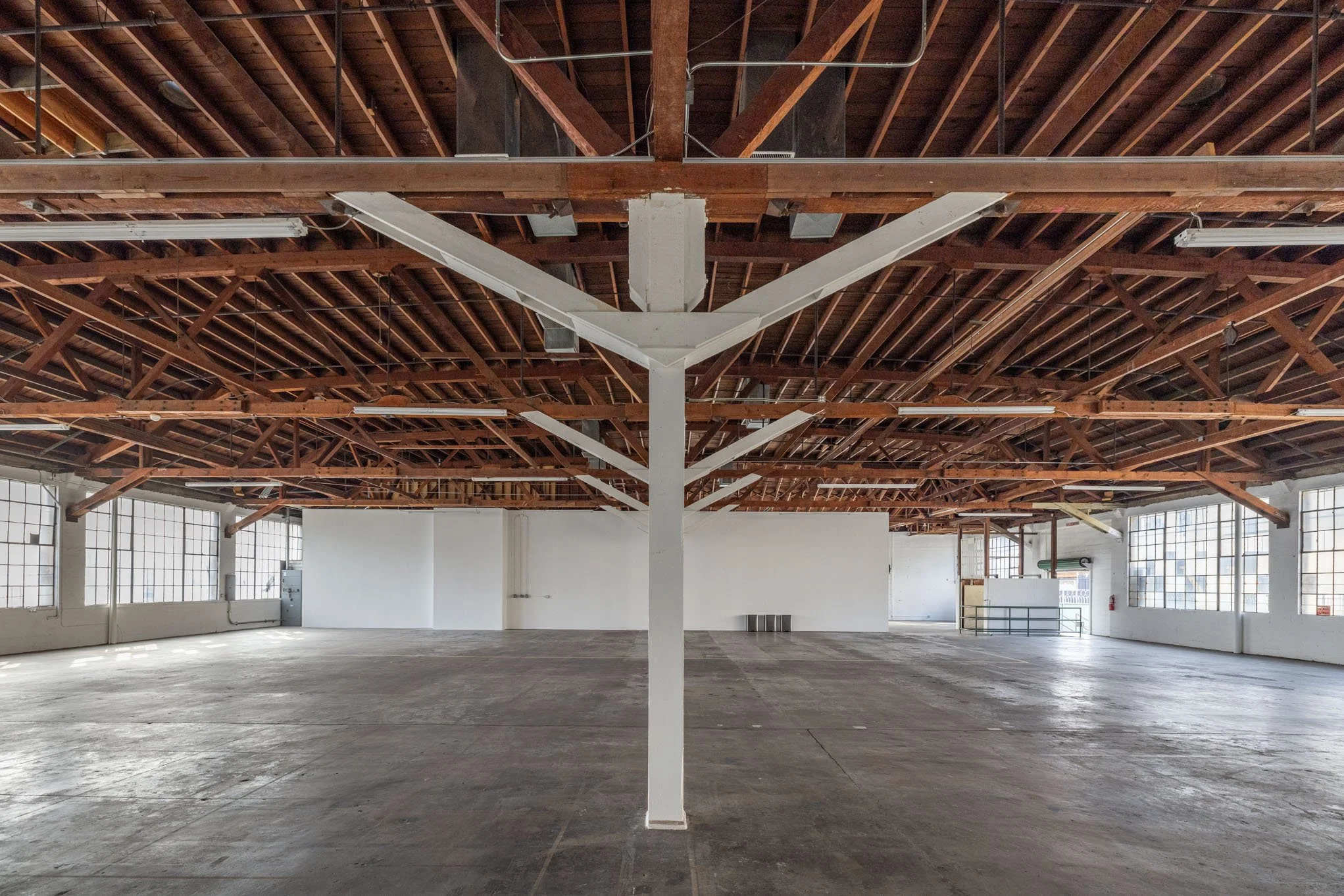 Empty industrial warehouse with wooden beam ceiling, large windows, and concrete floor.