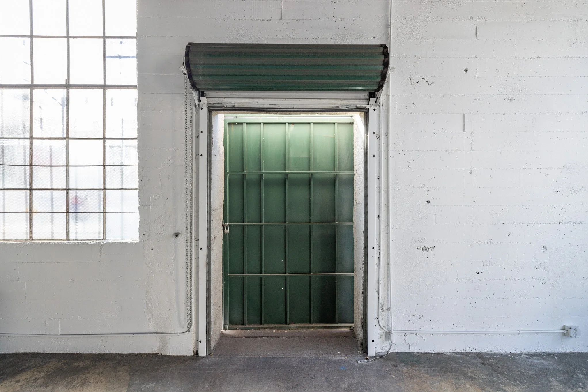 Interior of an industrial space with white painted brick walls, a green metal roll-up door, and large industrial window with grid pattern on the left.