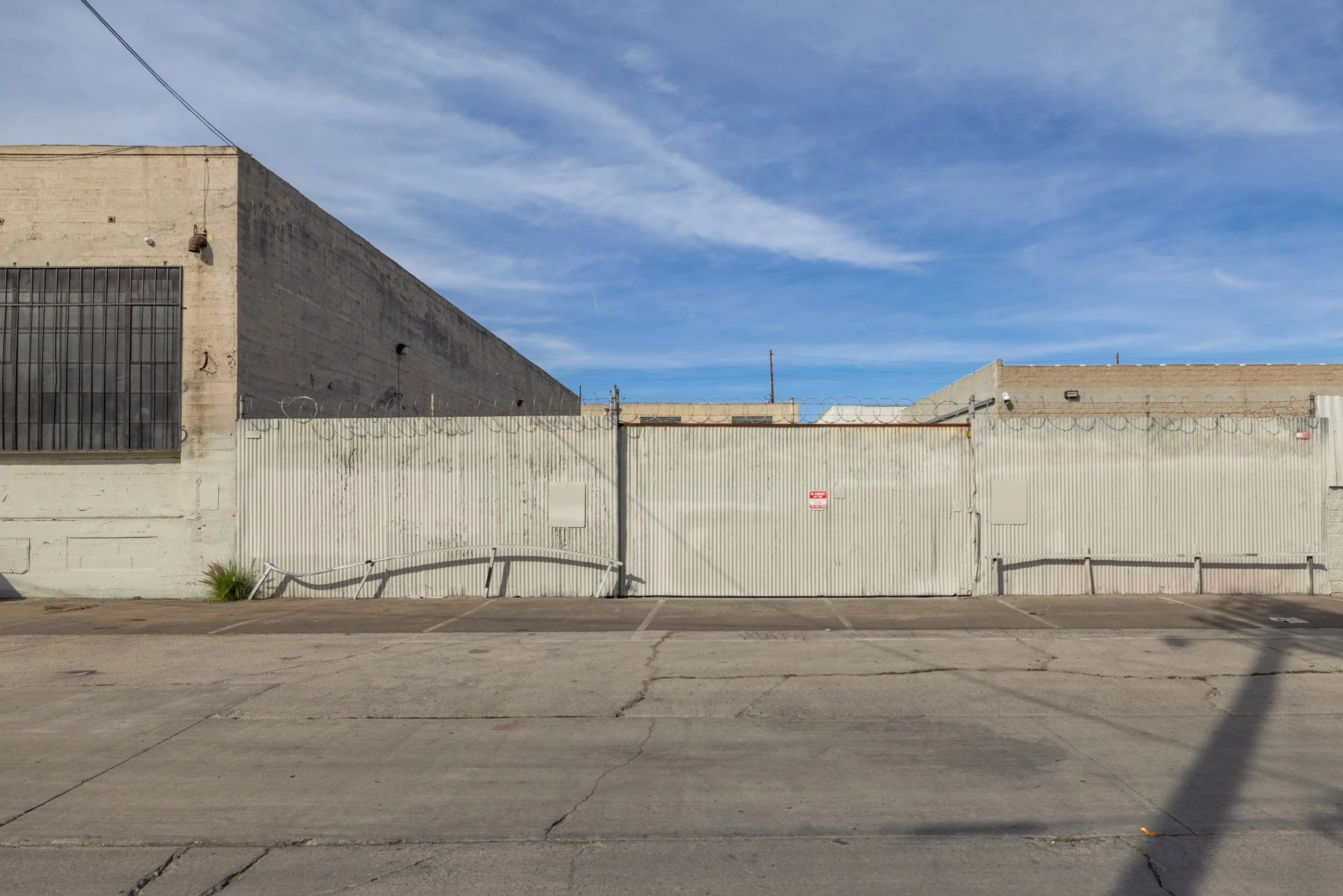 A chain-link fence topped with razor wire secures a warehouse or industrial building, with a cloudy blue sky overhead.