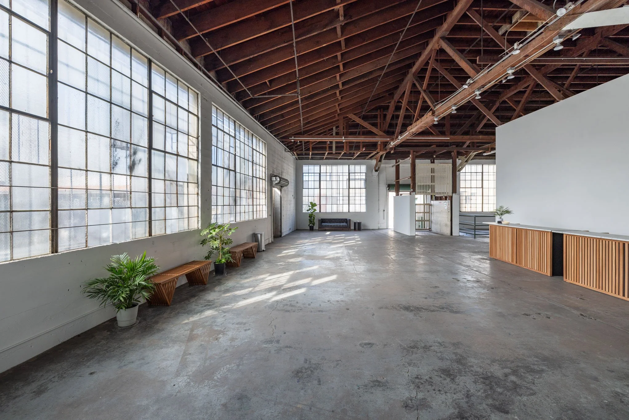 Spacious industrial-style room with large windows, wooden ceiling beams, concrete floor, potted plants, and minimalist furniture.
