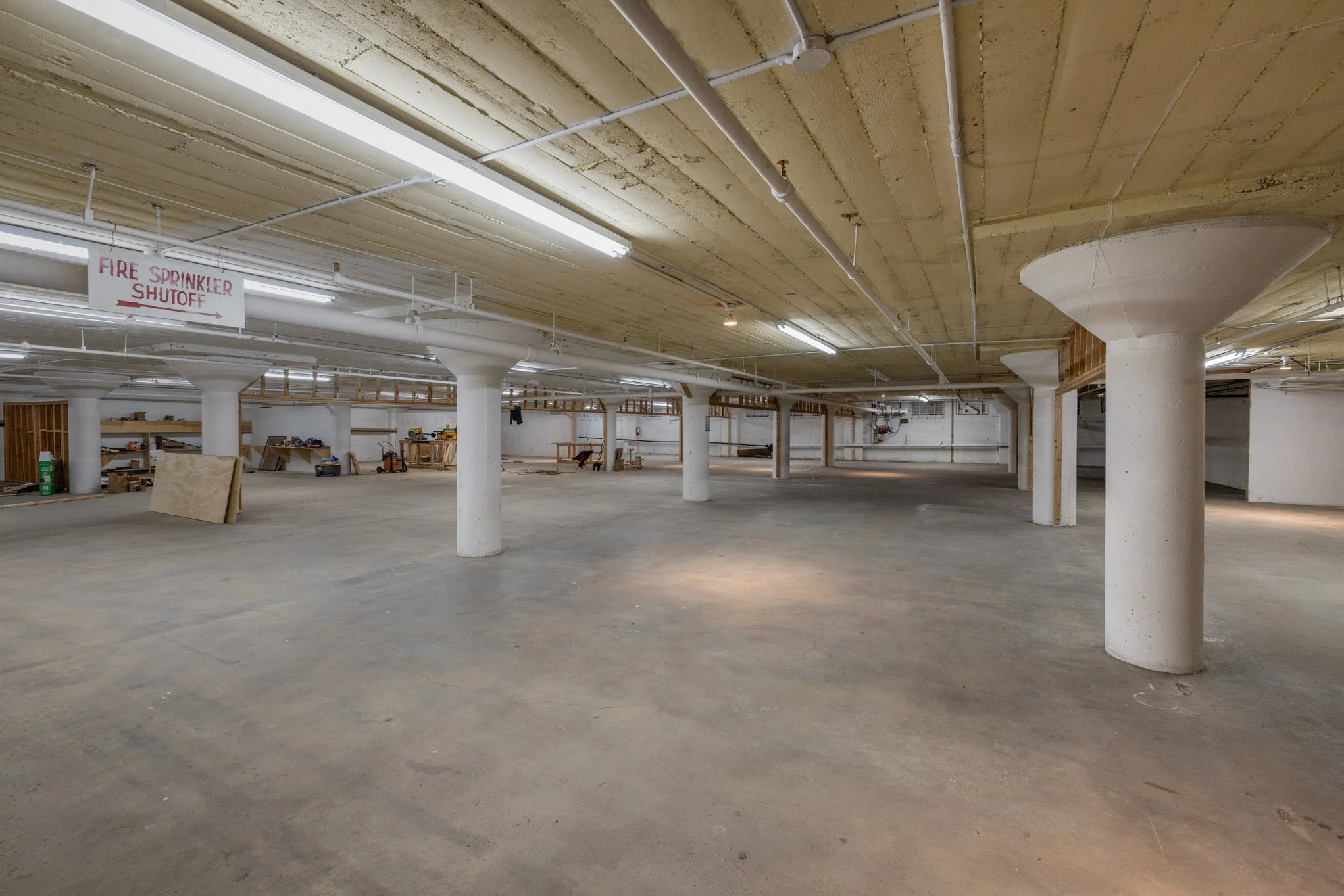 Empty parking garage with concrete pillars and fluorescent lighting, construction materials, and a sign indicating fire sprinkler shutoff.