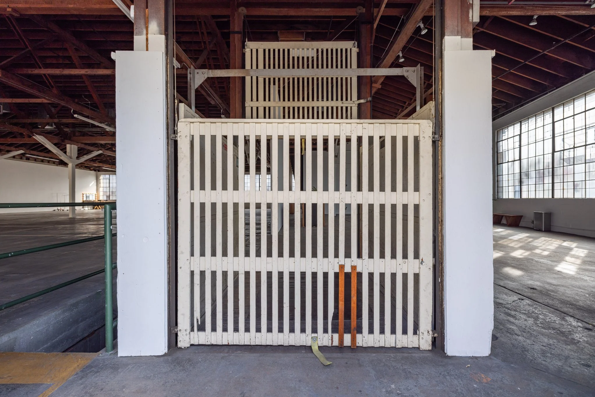 Indoor empty basketball court with a cage gate at the entrance, high ceilings, large windows, and wooden roof beams.