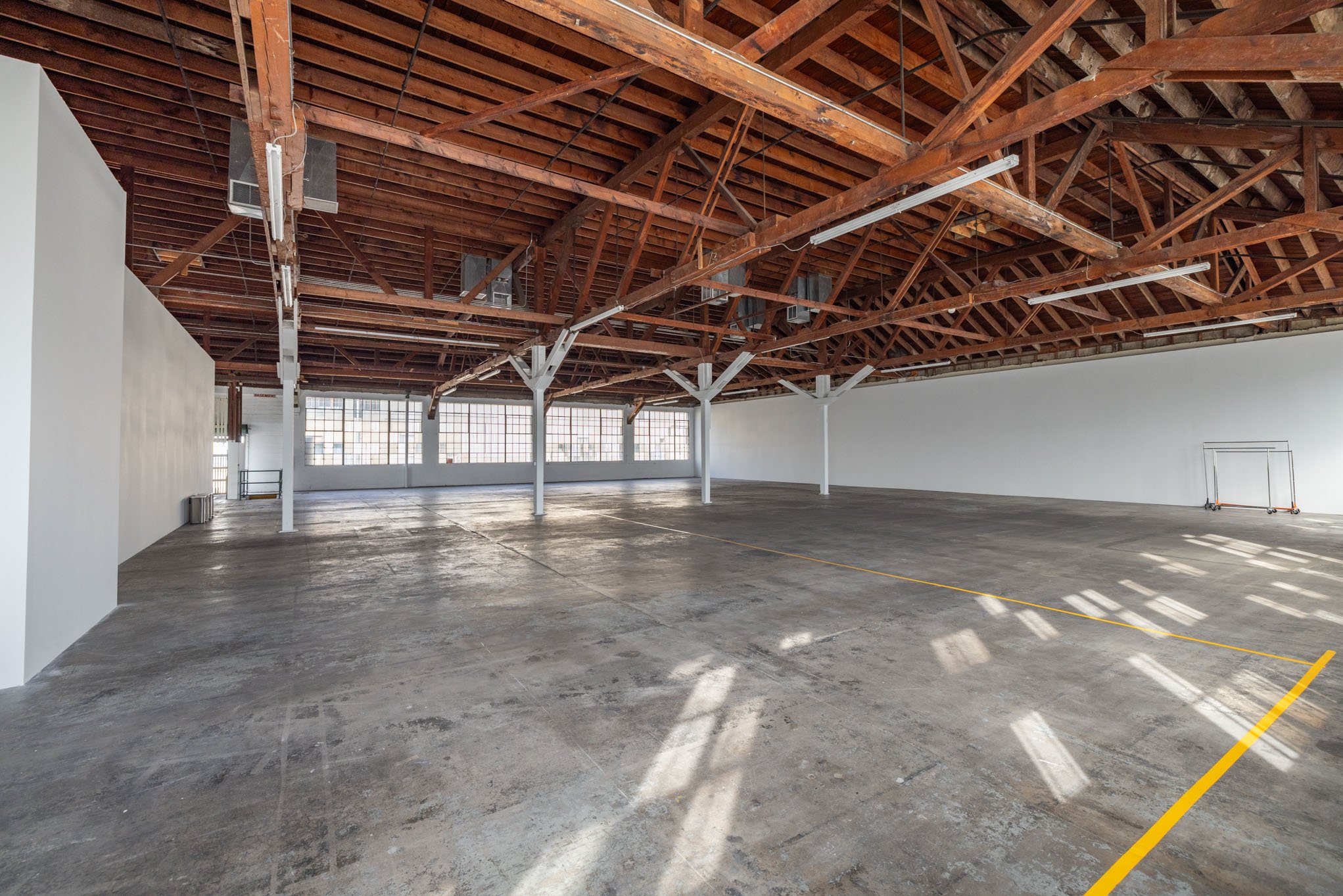 Empty industrial loft with concrete floor and exposed wooden ceiling with beams, large windows, and white support columns.