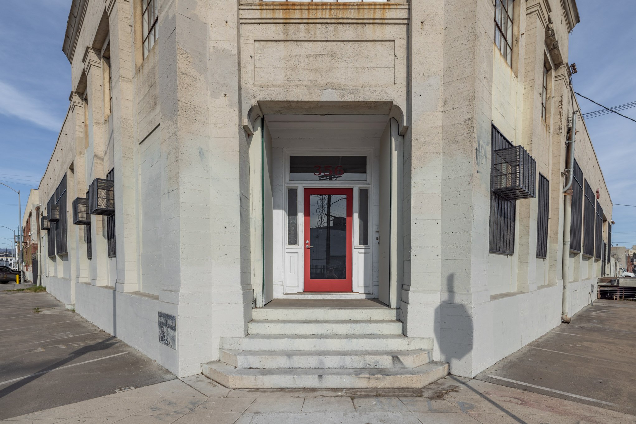 Corner of a white brick building with steps leading to a glass door framed in red, surrounded by windows with black security bars, under a blue sky.