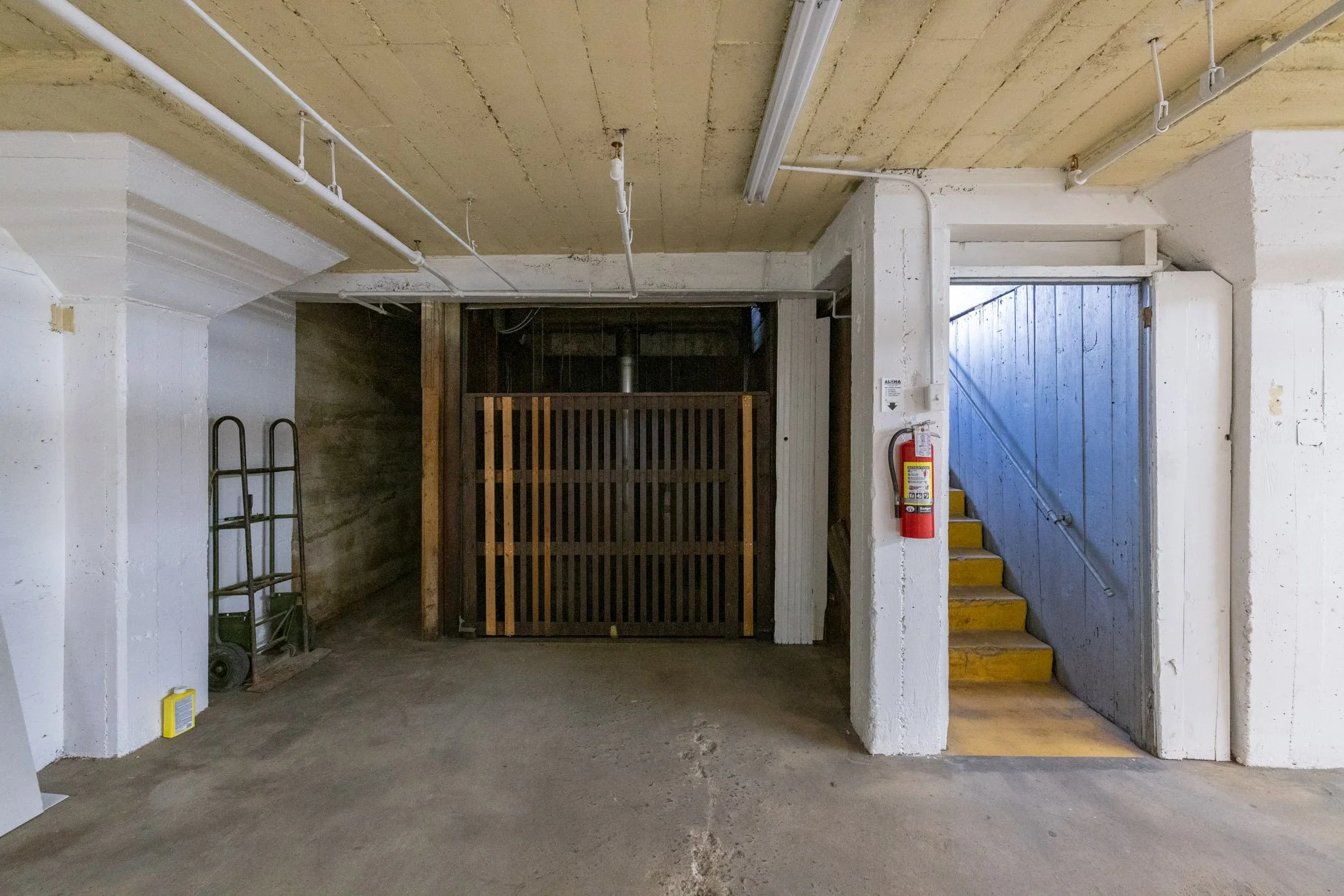 Underground parking garage with concrete flooring, white walls, wooden staircase with yellow steps, fire extinguisher on wall, metal cart, and ladder.