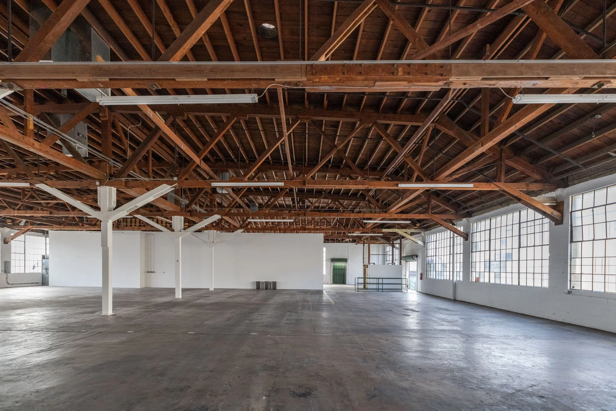Empty industrial warehouse with high wooden ceiling beams, large windows on the right, and a concrete floor.