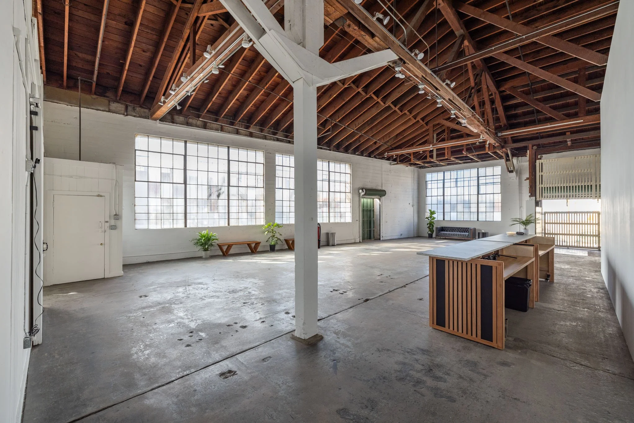 Spacious industrial-style room with large windows, exposed wooden beams, minimal furniture, and a concrete floor.