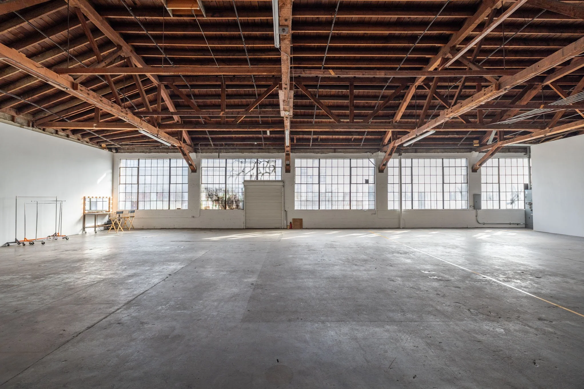 Empty industrial warehouse with large grid windows, exposed wooden ceiling beams, and concrete floor.