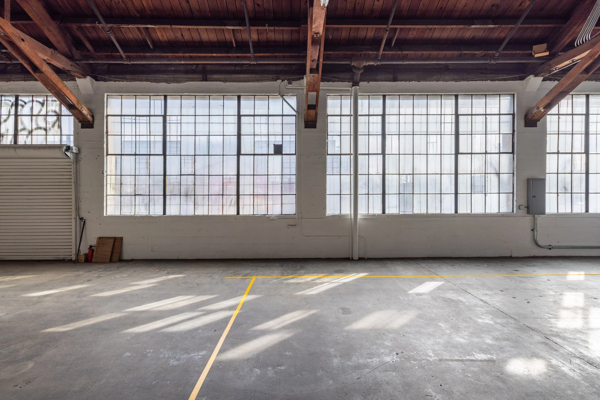 Empty industrial warehouse with large grid windows, concrete floor with yellow lines, scaffolding, and exposed wooden ceiling beams.
