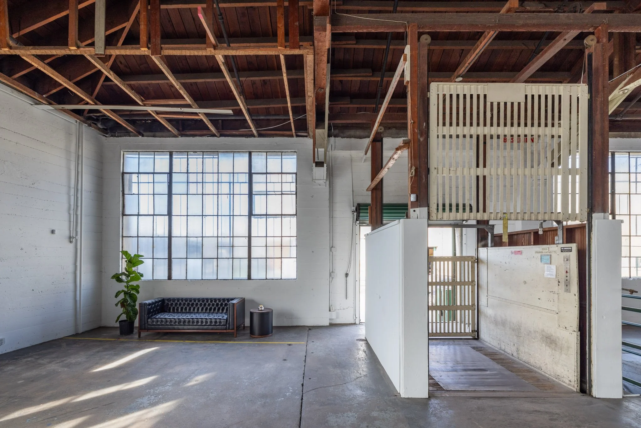 Industrial warehouse interior with large grid window, black leather couch, green potted plant, small black side table, metal and wooden structures, and an elevator or lift area.