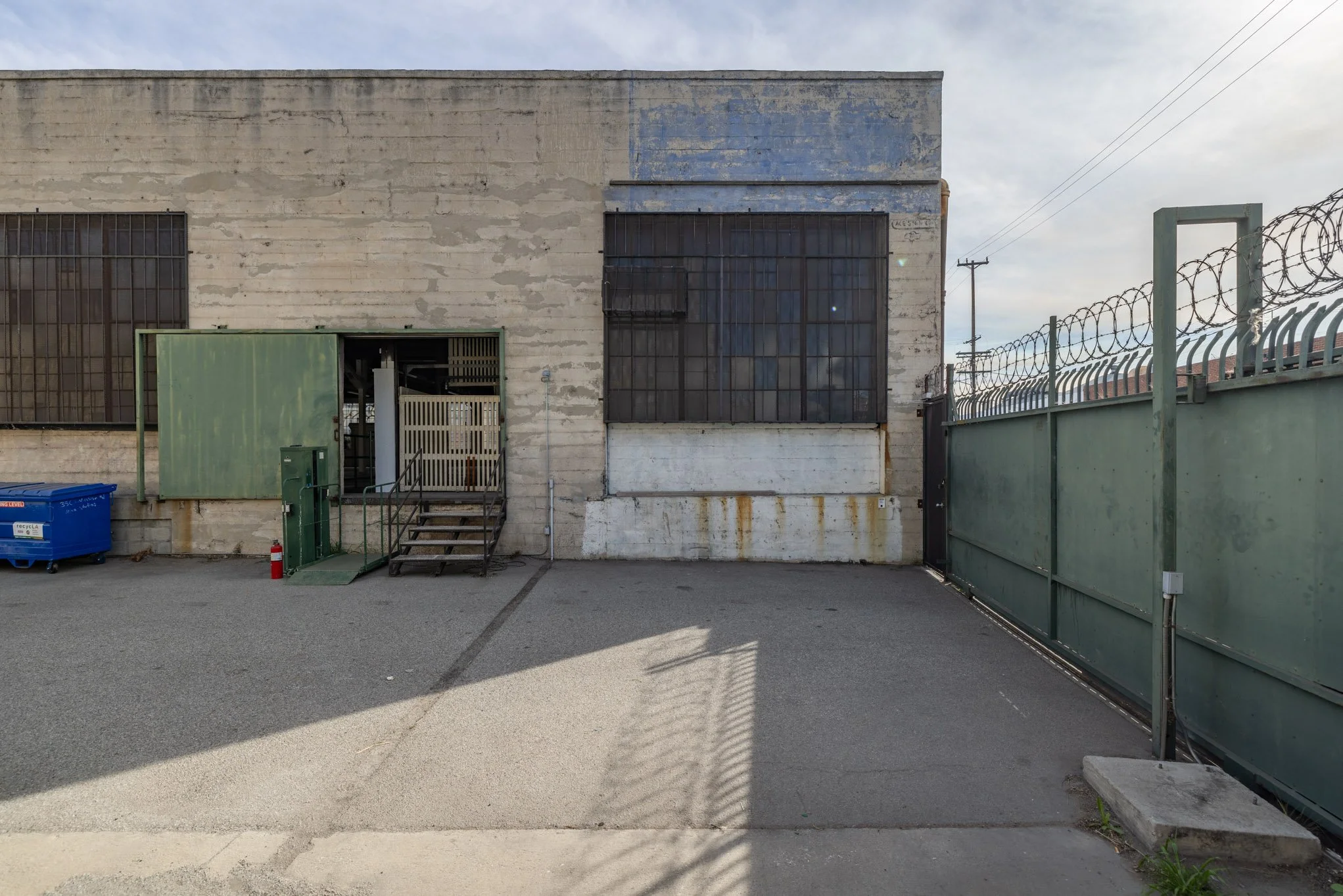 A plain, worn concrete building with barred windows, a green metal door, stairs, and a large green gate topped with barbed wire, in an outdoor lot.