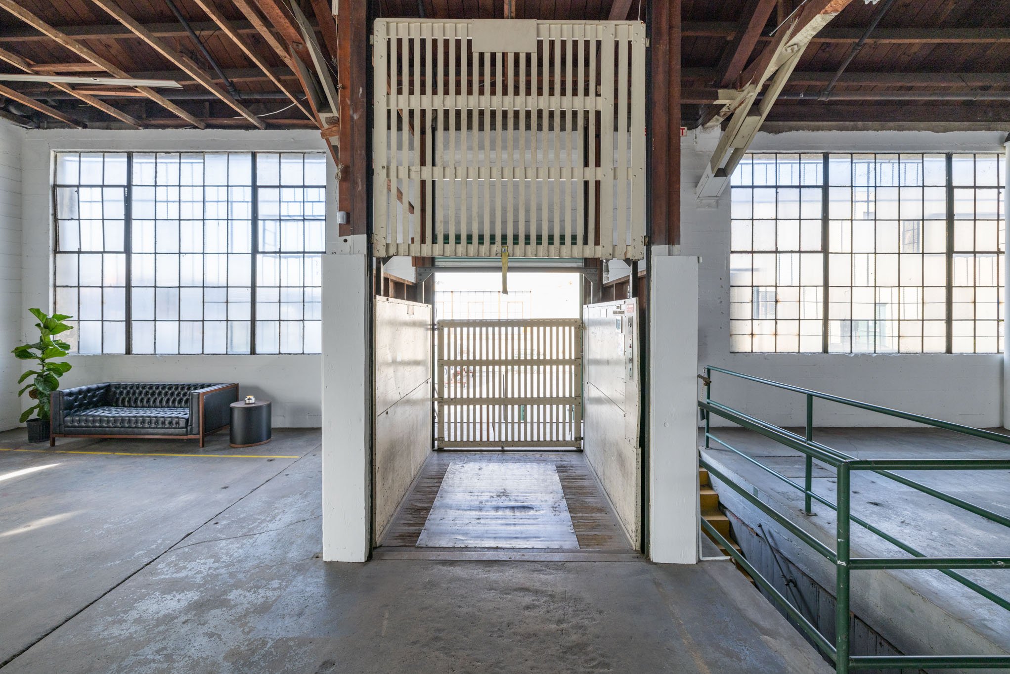 Industrial warehouse interior with large windows, a black tufted leather sofa with a small side table, a potted plant, and a vintage elevator with wooden panels and gated door at center.
