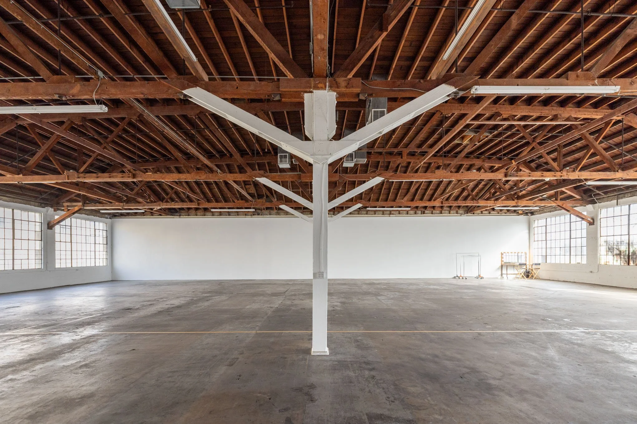 Empty industrial-style warehouse with a concrete floor, large windows, exposed wooden ceiling beams, and a white support column in the center.