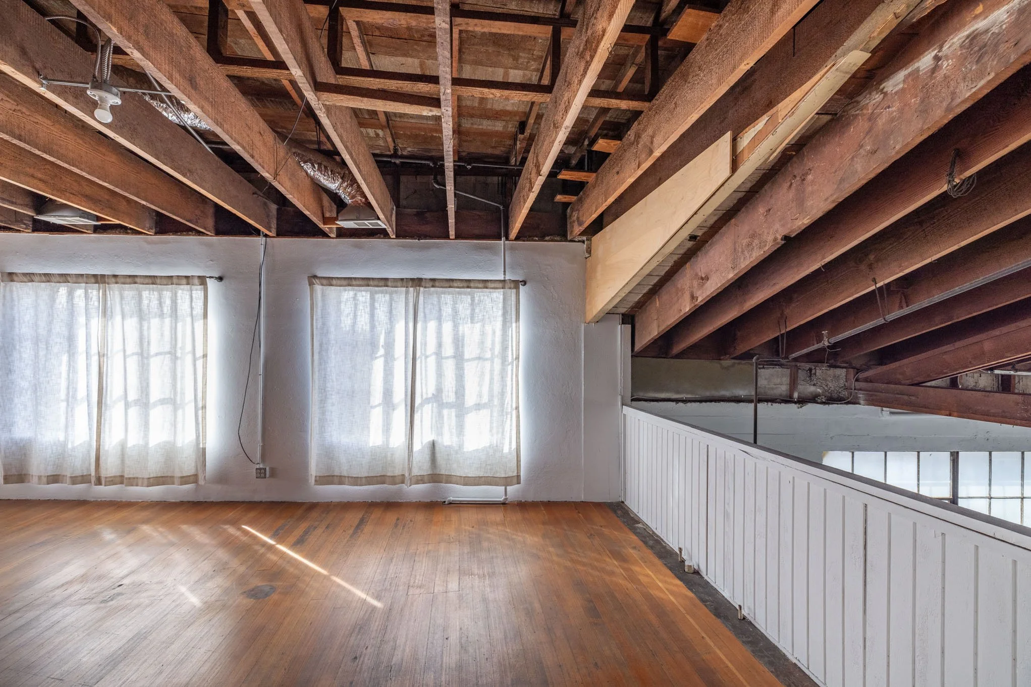 Empty interior space with wooden floor, white wall with two window curtains, exposed ceiling beams, and a partial railing.